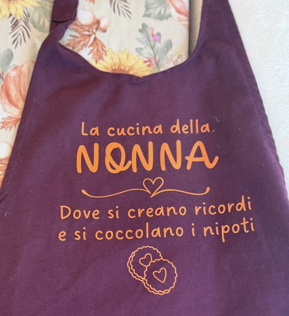 Purple apron with orange text about a grandmother's kitchen, featuring a small heart and flower design, on a floral background.