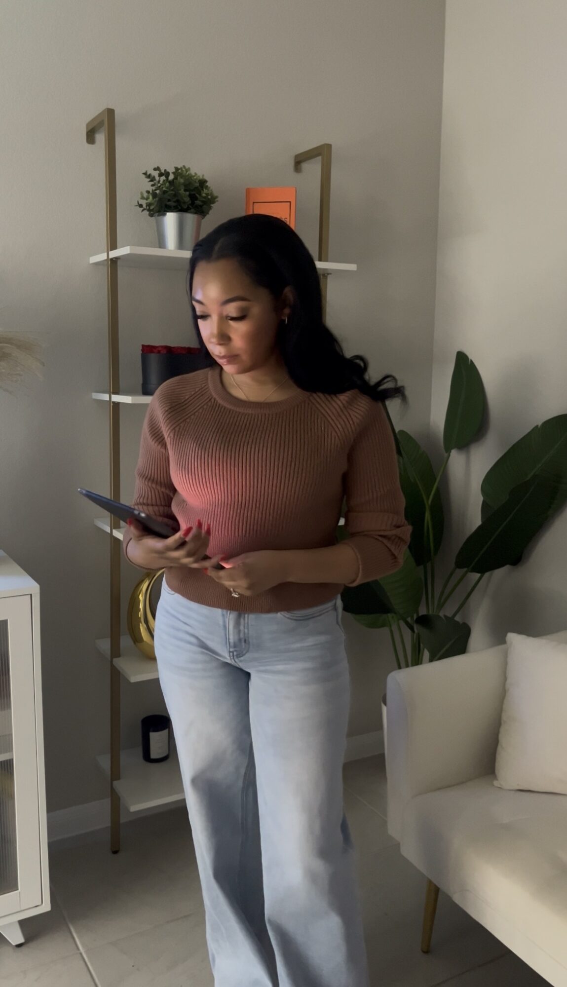 Woman with long dark hair wearing a brown sweater and light jeans, looking at her phone in a living room.
