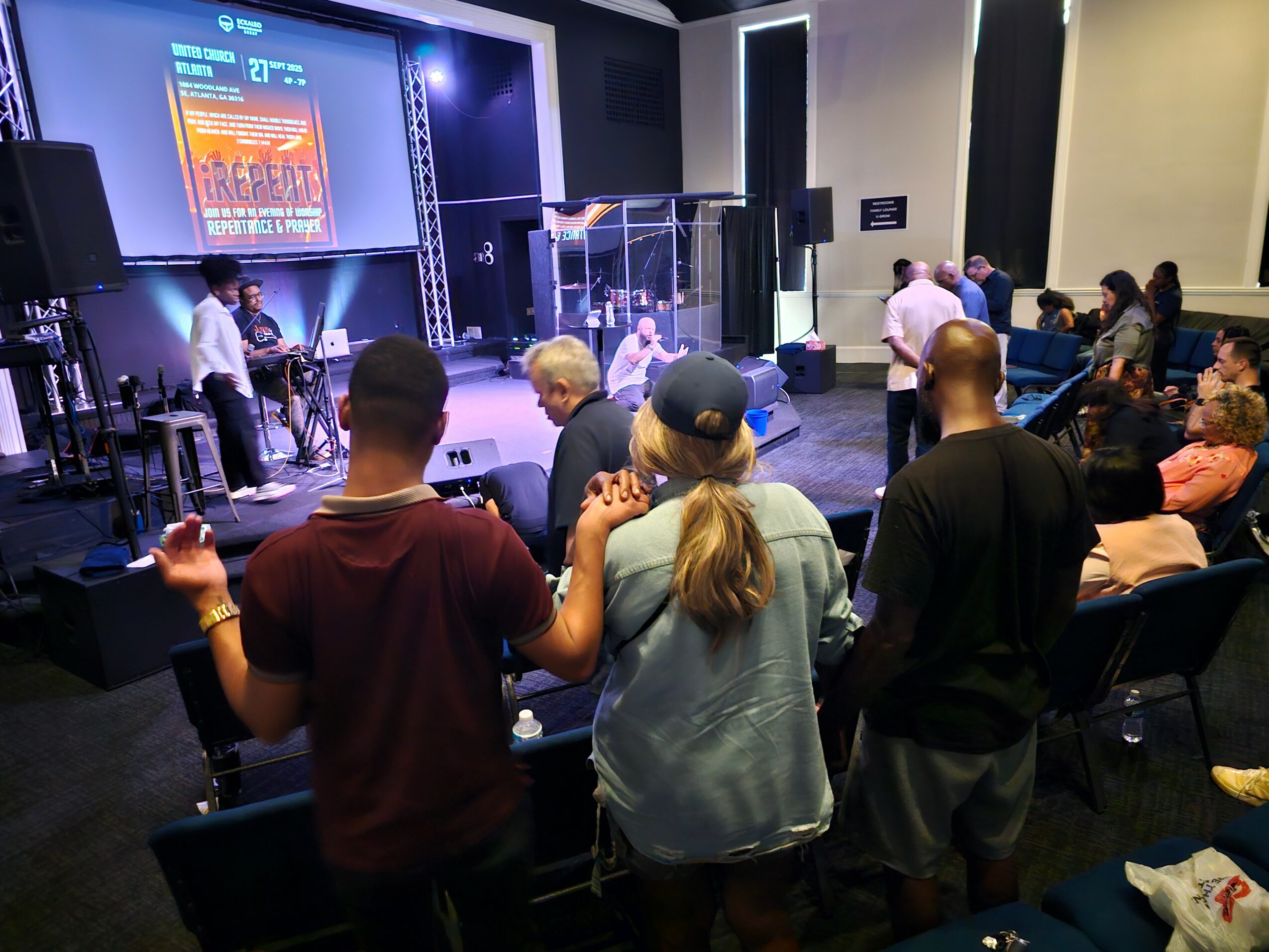 People gather in a church or conference room with a stage, screen, and musical instruments, attending a service or event.