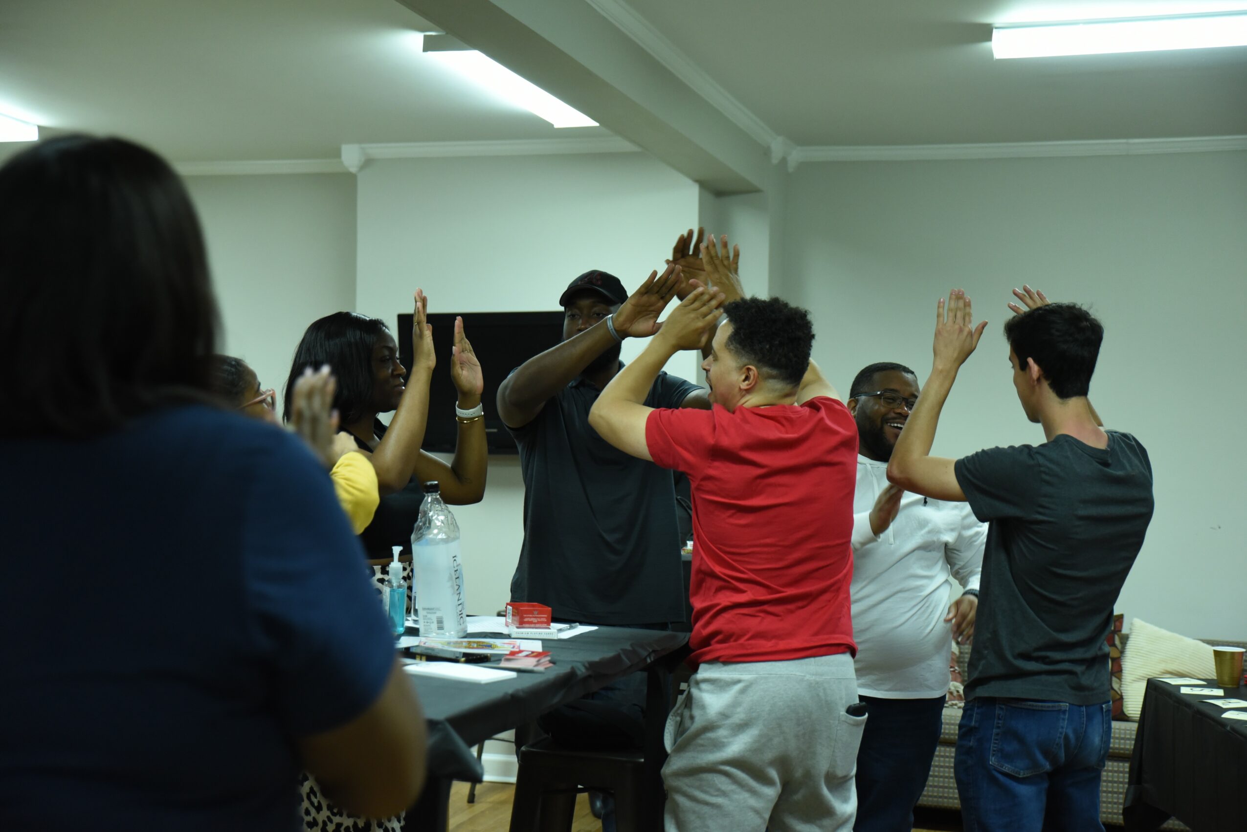 Group of people in a room giving high-fives, some facing each other, with a table and bottles in the background.