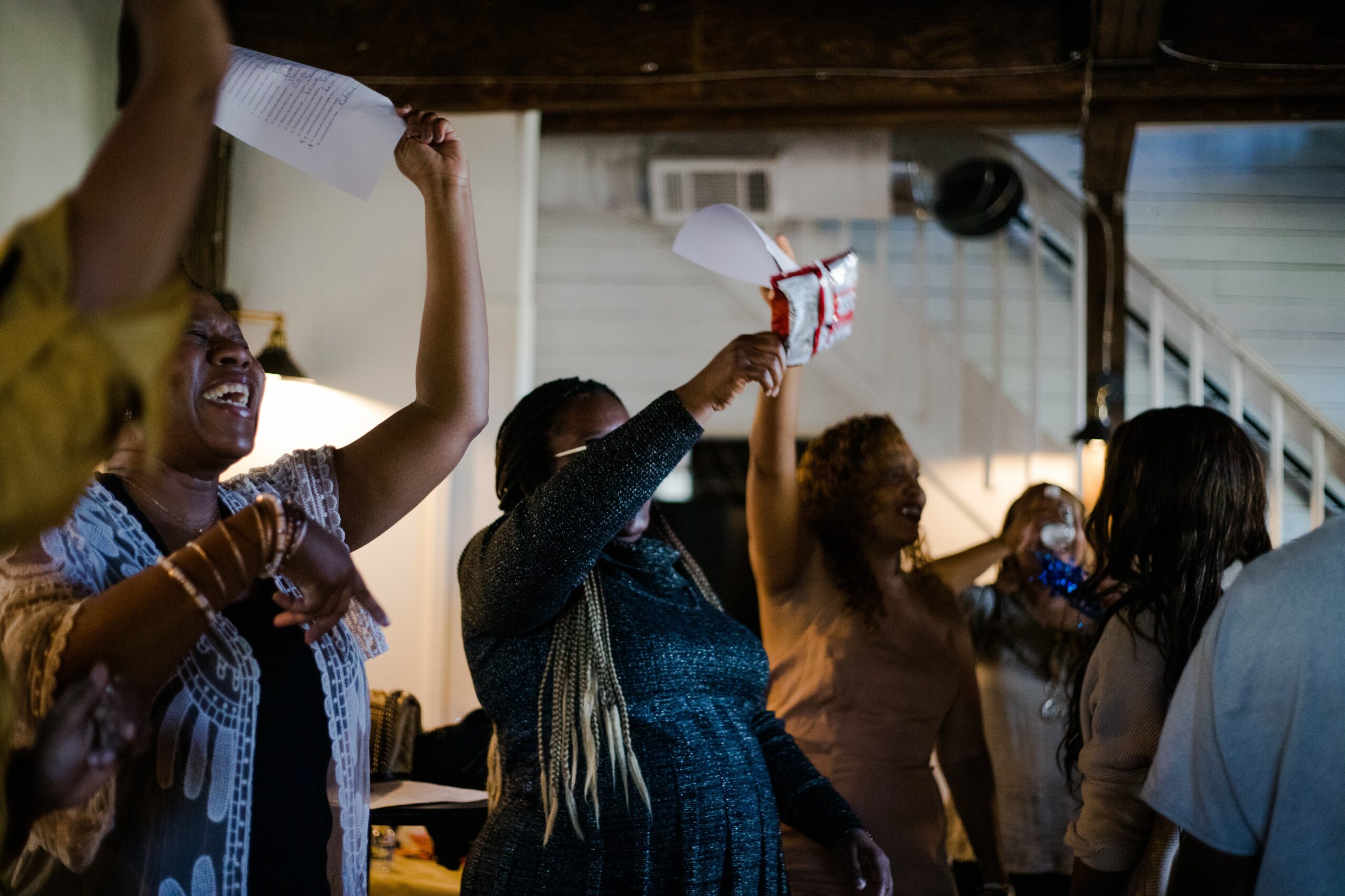 Group of women celebrating indoors, holding papers and raising hands, some wearing masks, with a wooden ceiling and window in background.