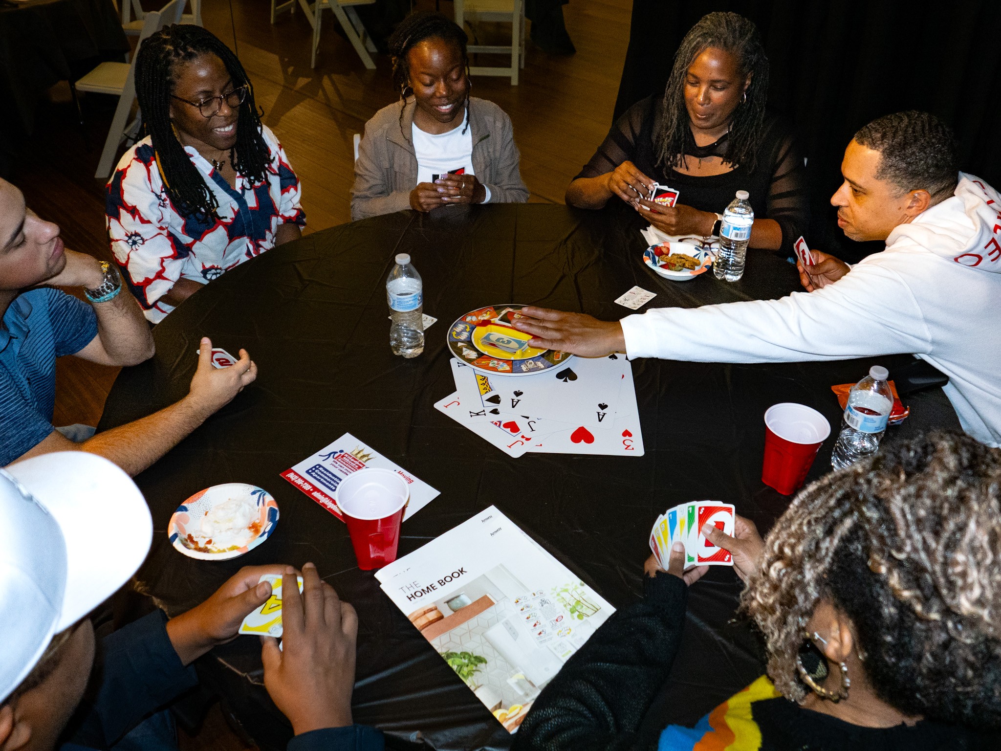 Group of people playing cards around a table with drinks and snacks, smiling and engaging in game.
