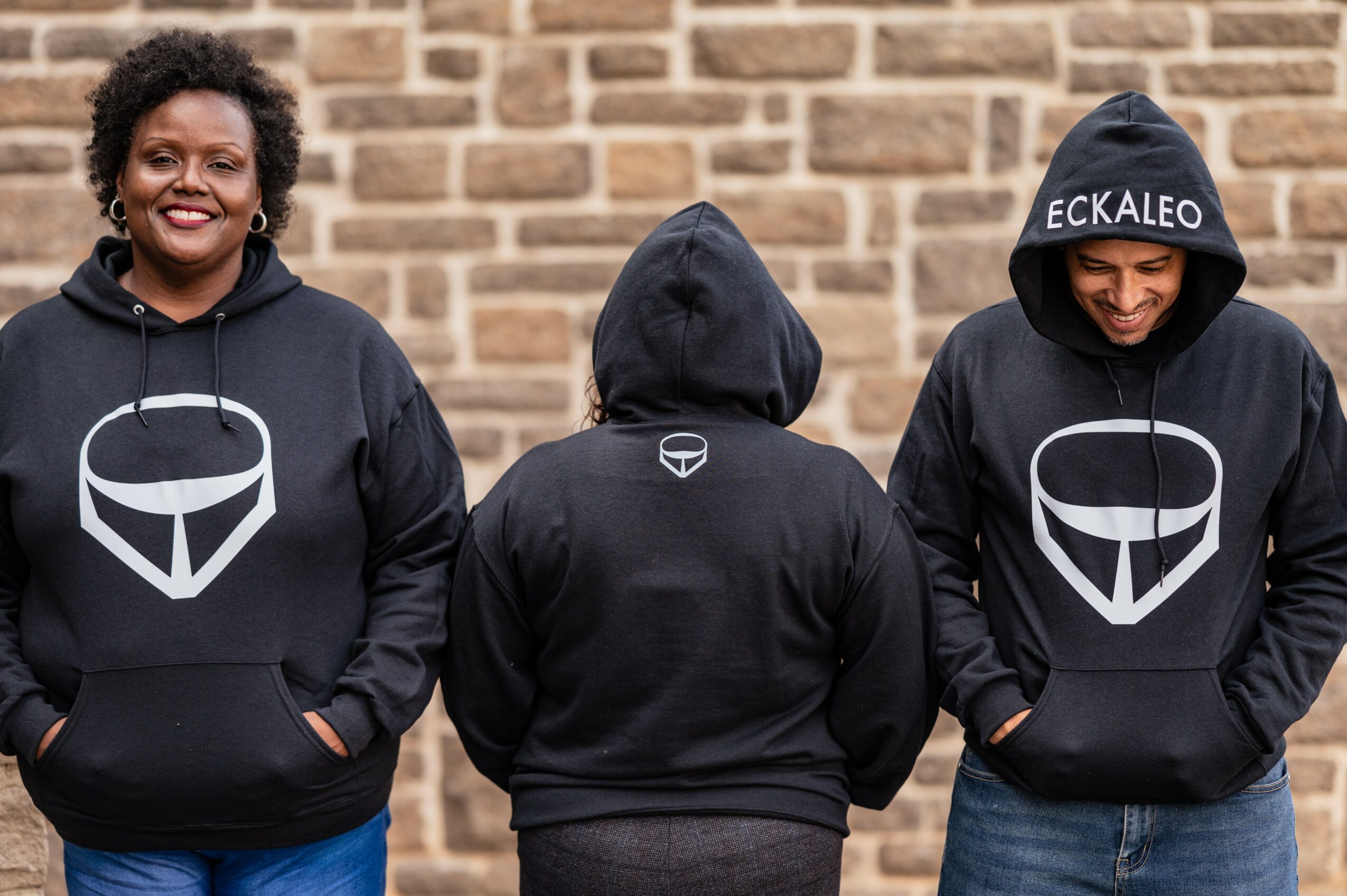 Three people wearing black hoodies with a white logo, standing against a brick wall, two facing forward and one with back to camera.