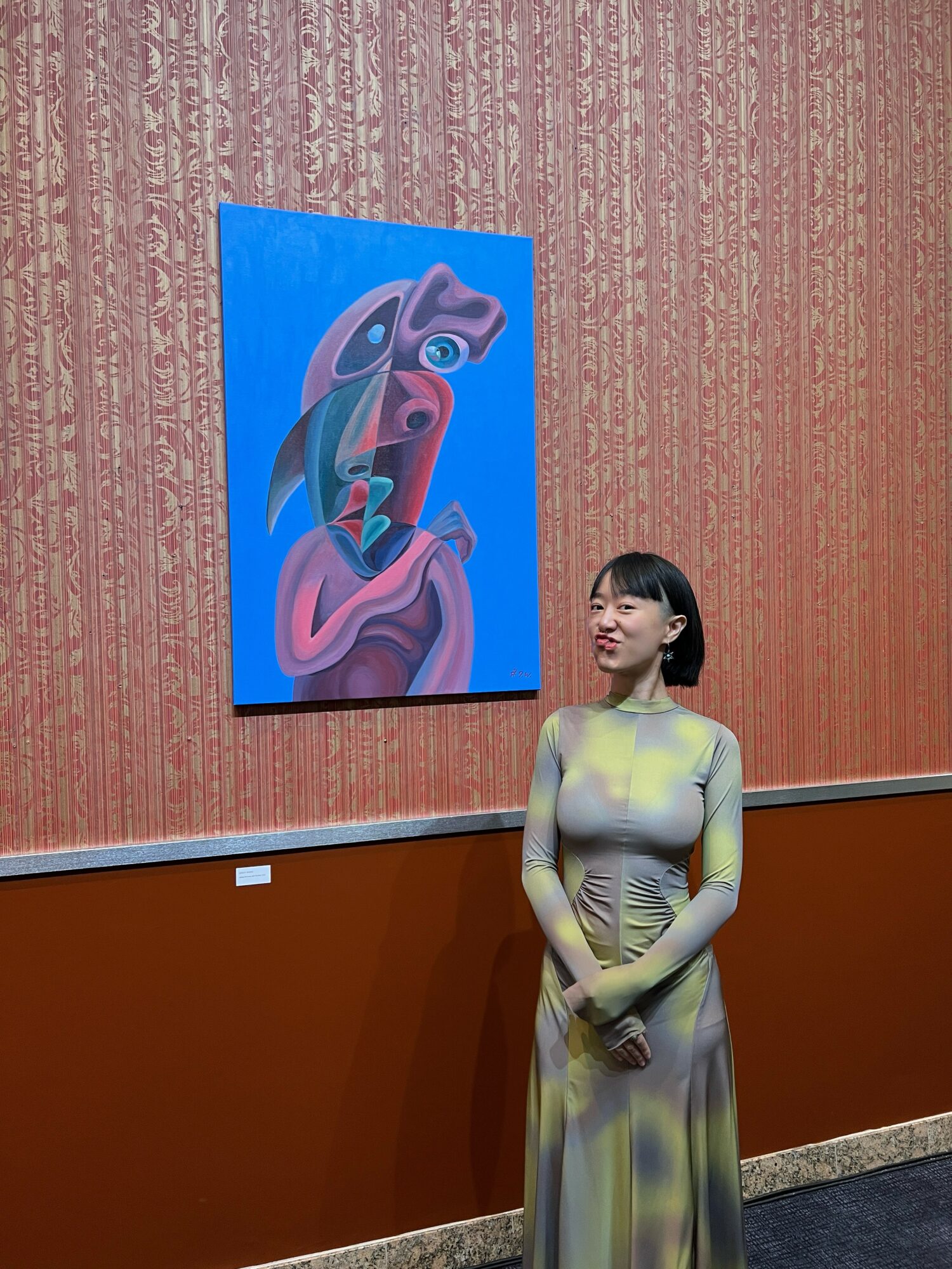 Young woman in a long, patterned dress stands in an art gallery, looking at a colorful abstract painting on the wall.