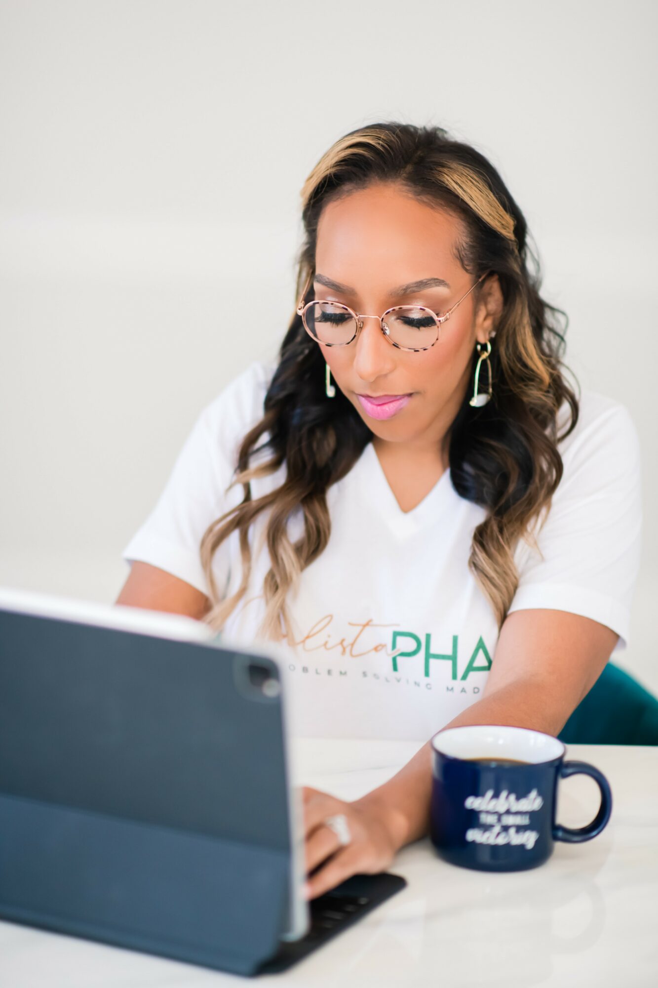 Woman with glasses and earrings working on a laptop, with a mug nearby, against a plain background.