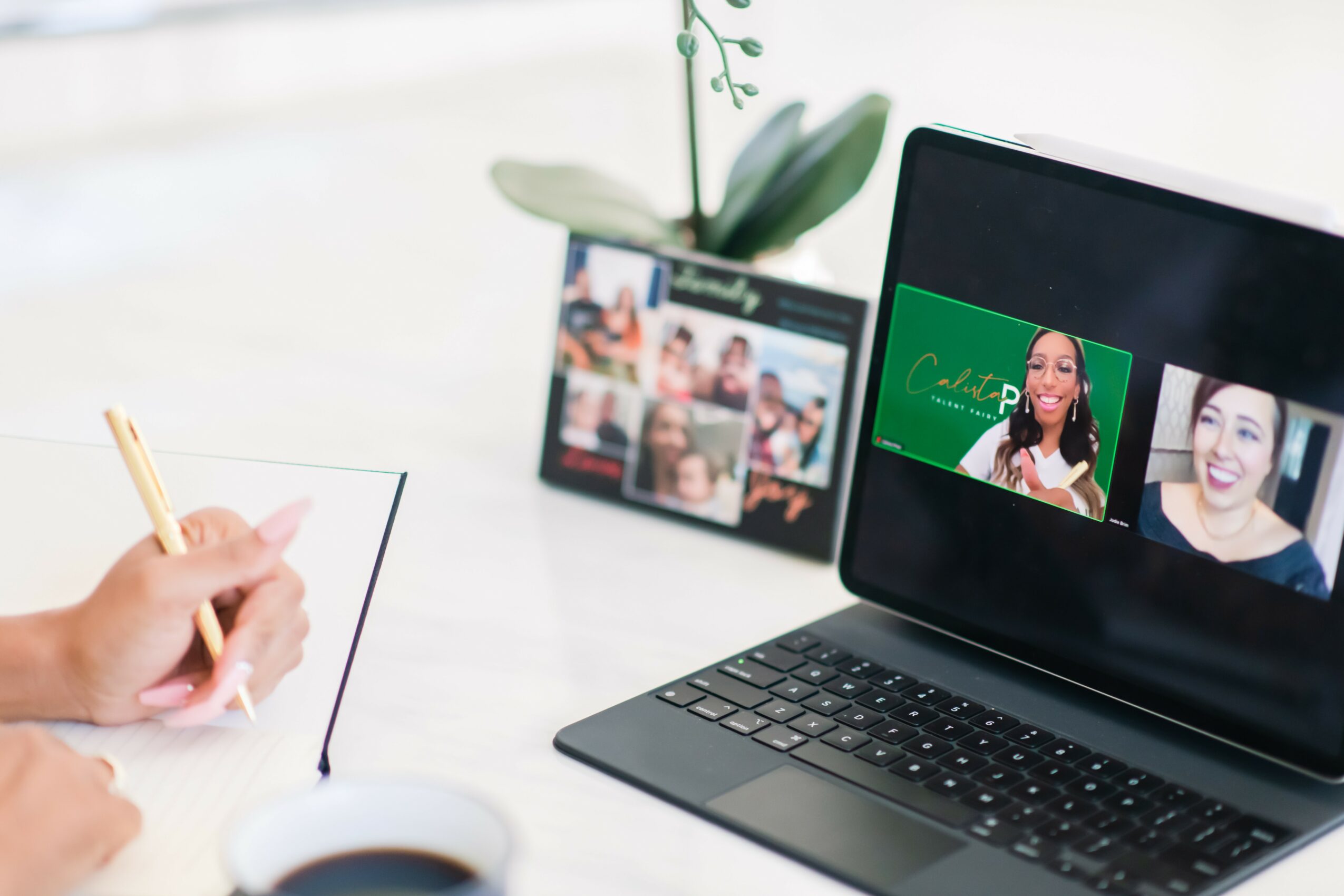 Person taking notes during a virtual meeting on a laptop with two women smiling on screen, a tablet showing a video call, and a plant in the background.
