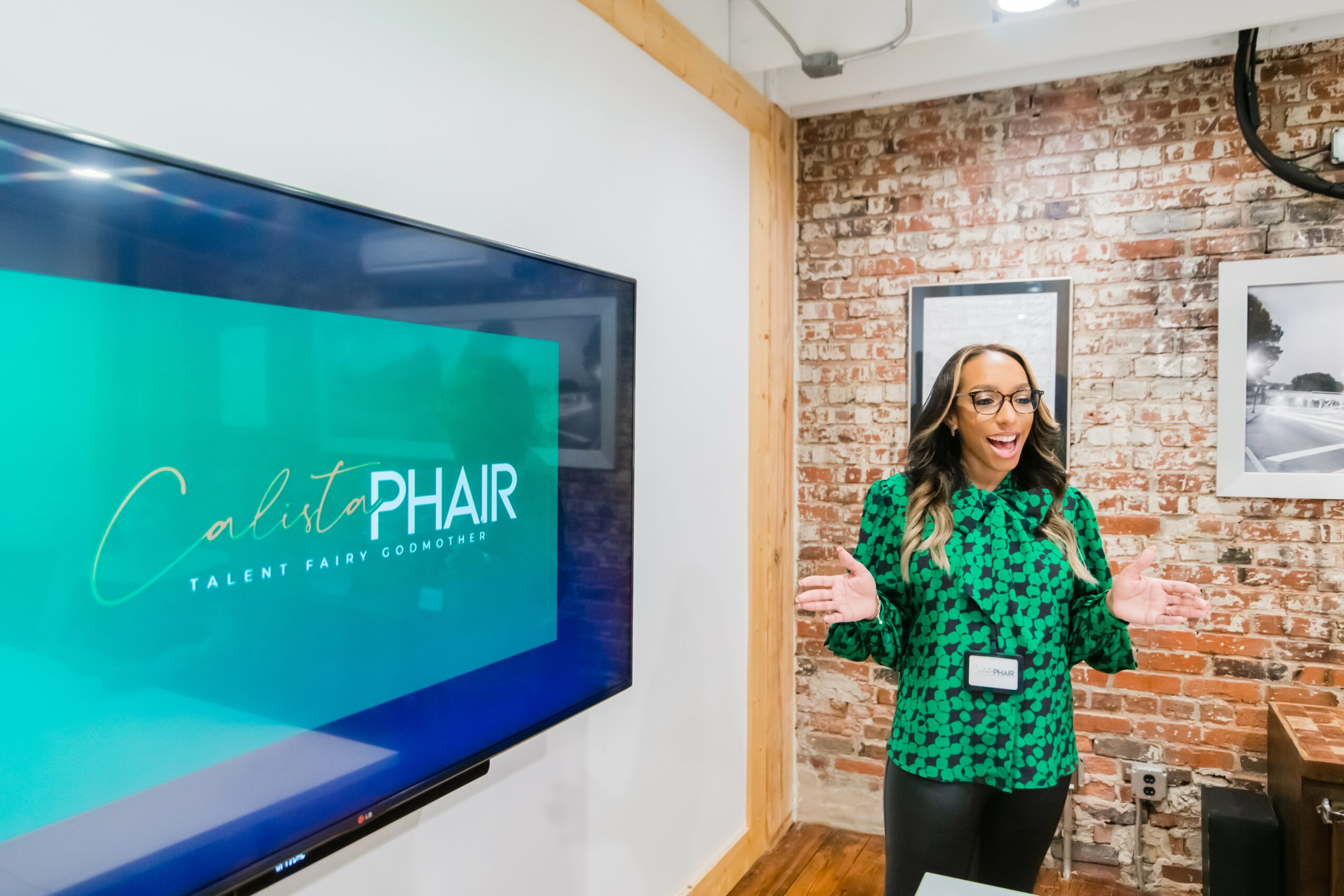 Woman with glasses and long hair speaking in a room with brick wall, large screen, and framed pictures.