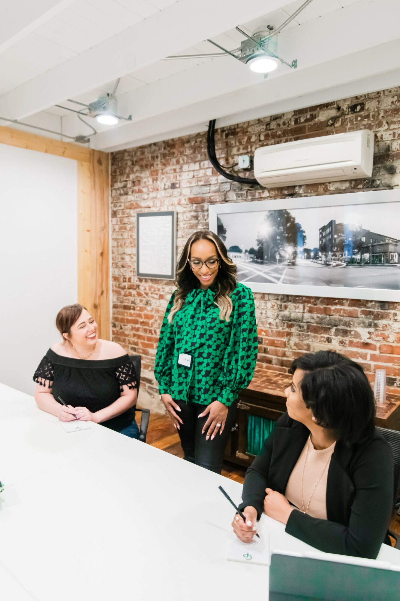 Three women in a room with brick wall, air conditioner, and large photo of a city street, engaged in conversation.