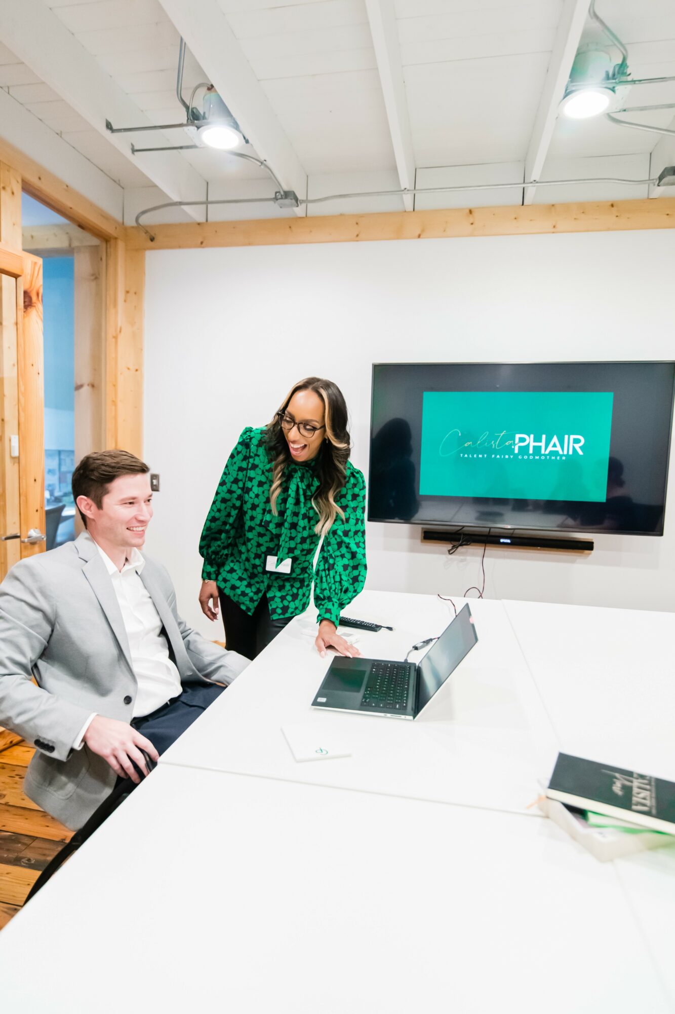 Two women in a meeting room with a large screen displaying 'PHAIR' and a laptop on the table.