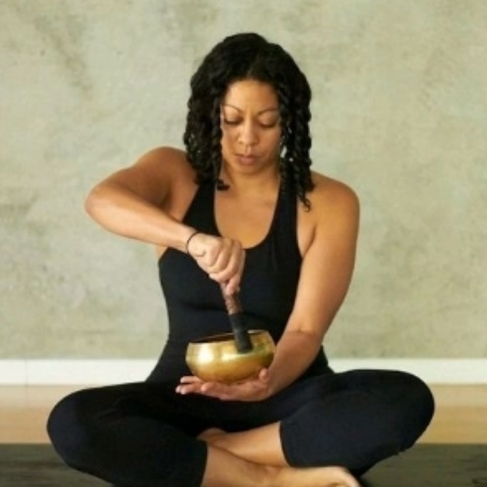 Woman sitting cross-legged, using a singing bowl with a mallet, against a plain background.