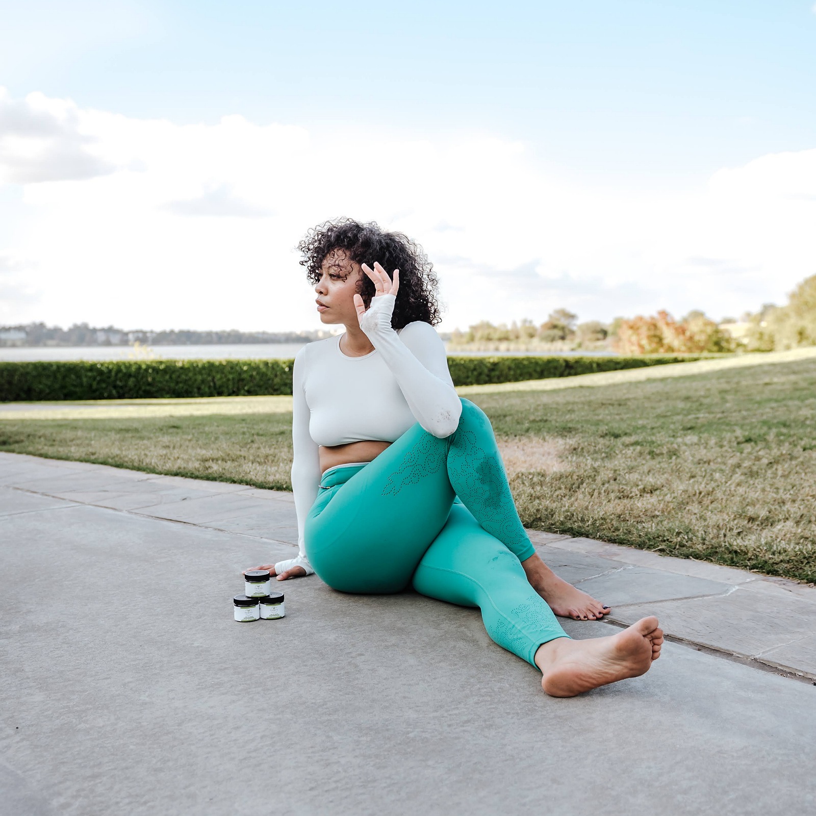 Woman sitting on sidewalk, talking on phone, wearing teal leggings and white top, outdoors with grass and sky.