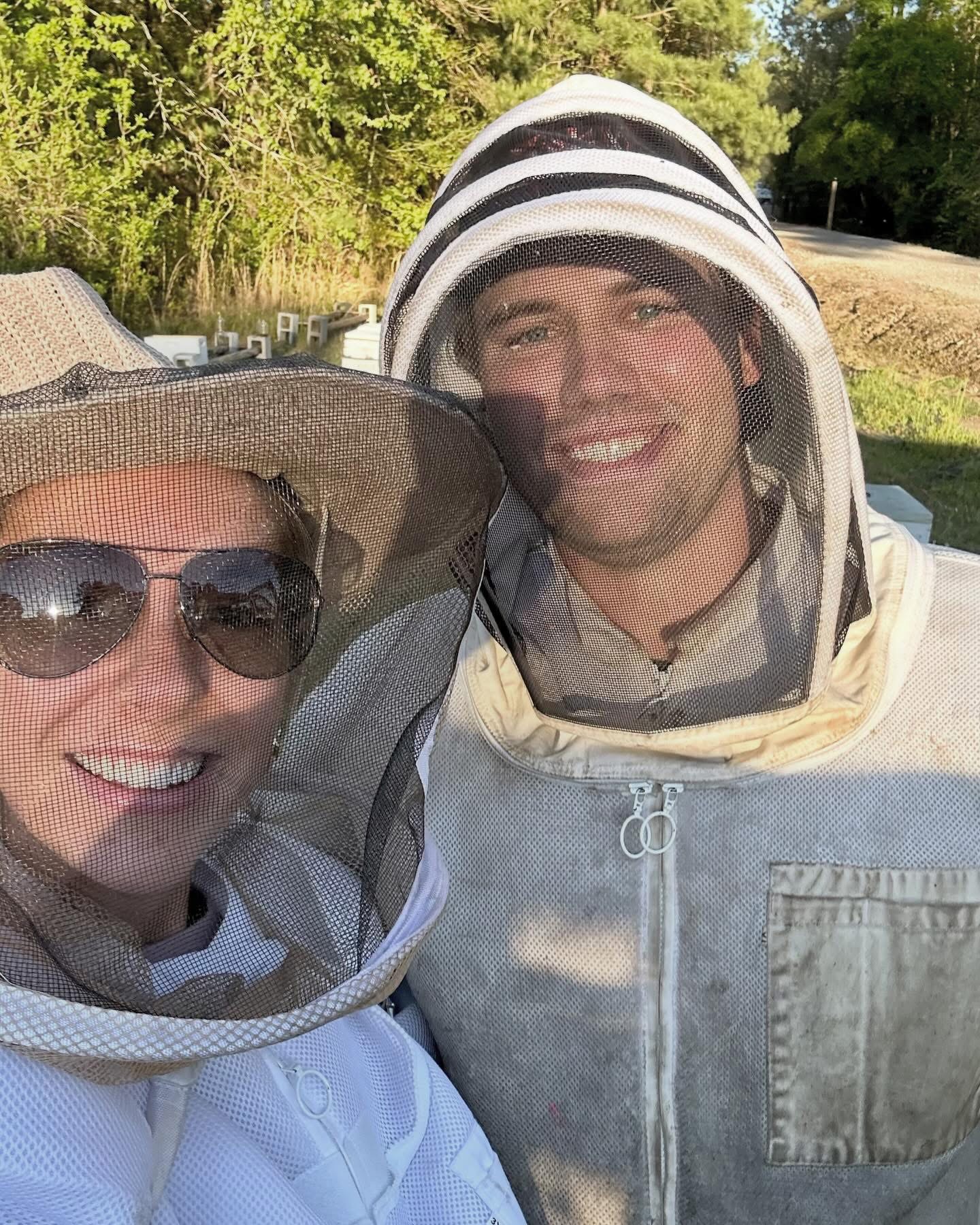Two people outdoors wearing protective hats and gear, smiling at the camera, with trees and a path in the background.