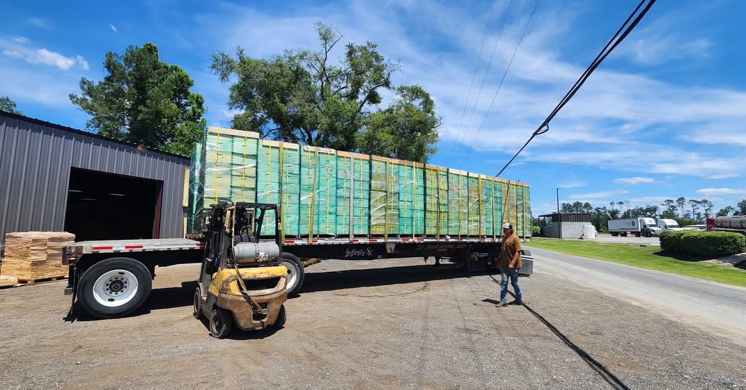 Flatbed truck loaded with stacked green and yellow crates, forklift nearby, person walking beside the truck, blue sky with clouds.