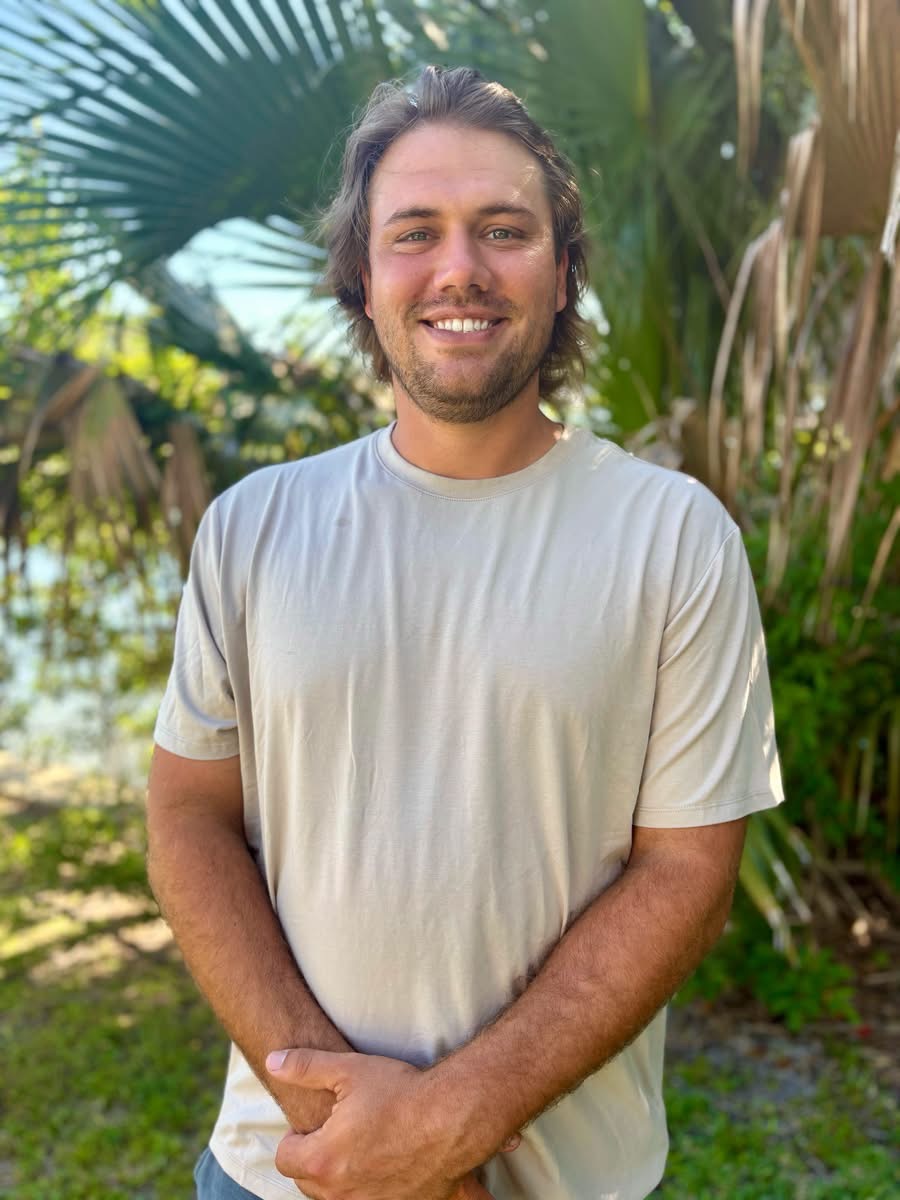 Smiling man with shoulder-length hair and beard standing outdoors among tropical plants.