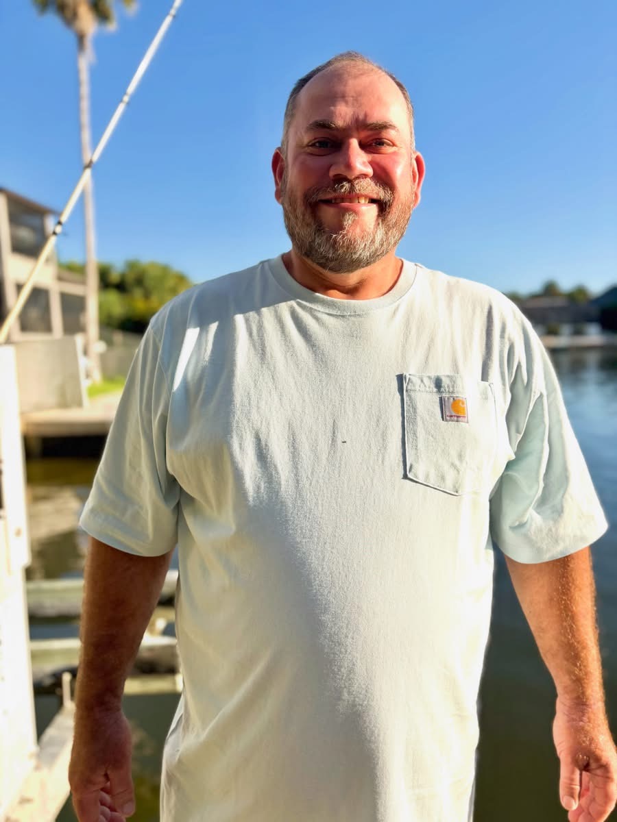 Man with a beard smiling outdoors near water, wearing a light-colored T-shirt with a pocket, sunny day.