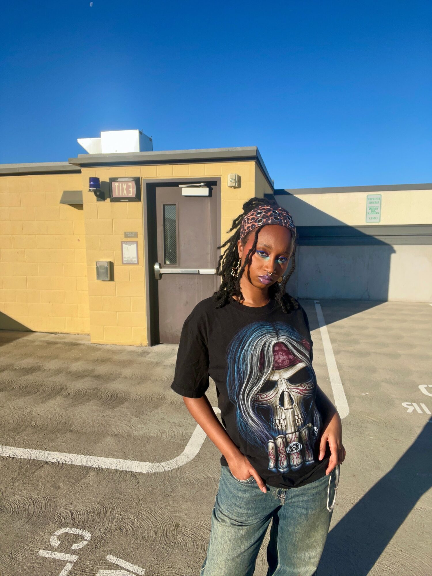 Young woman with dreadlocks and headband standing outdoors in a parking lot, wearing a black graphic t-shirt and jeans.