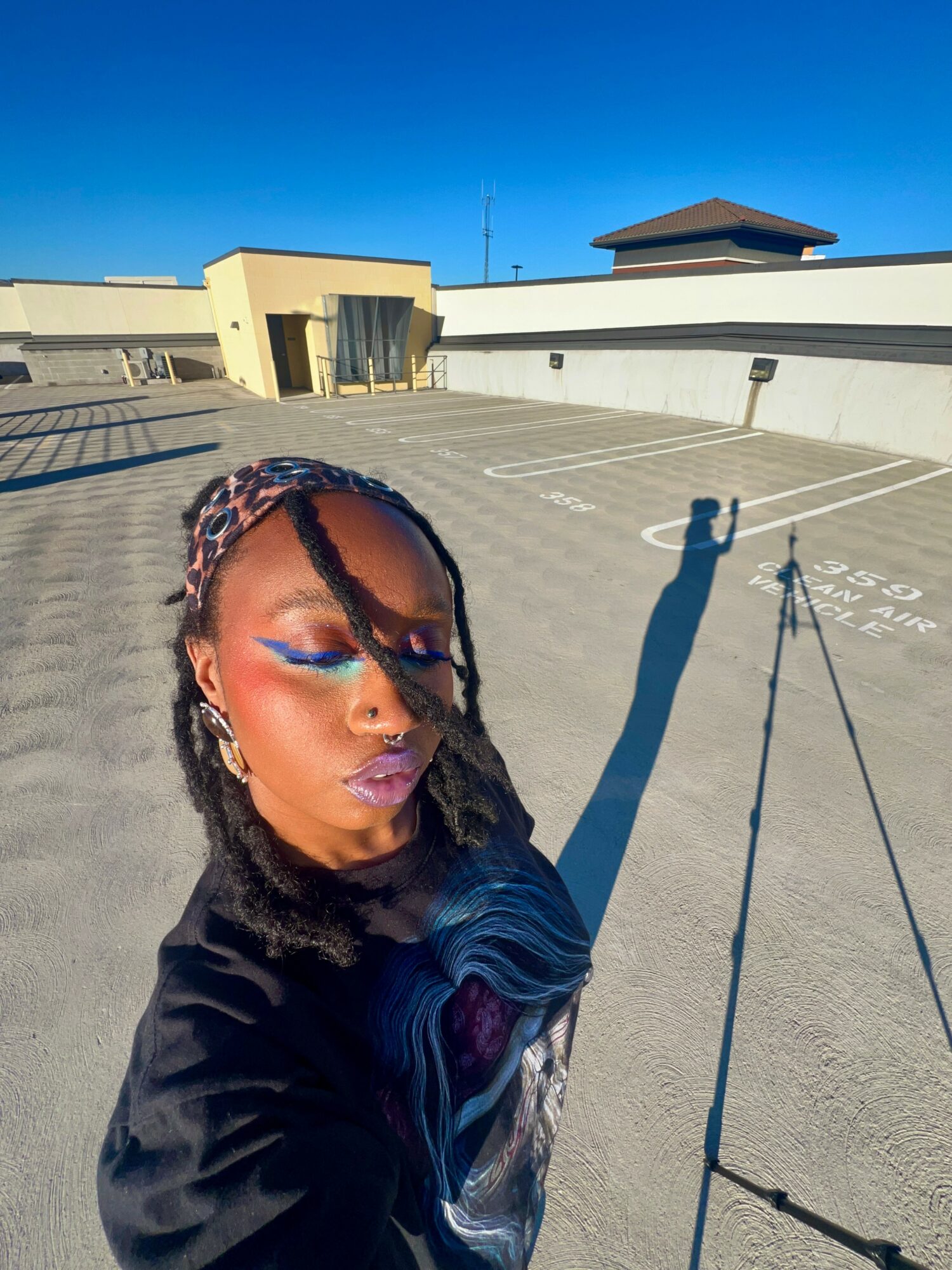 Person with colorful makeup and braided hair taking a selfie on a rooftop parking lot under a clear blue sky.