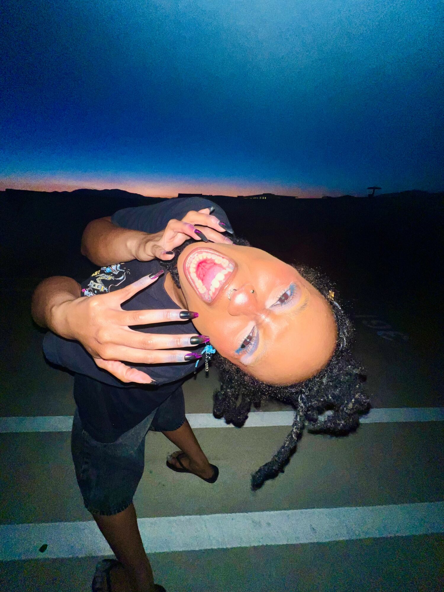 Person with curly hair and earrings smiling widely, making a playful gesture with hands, outdoors at dusk.