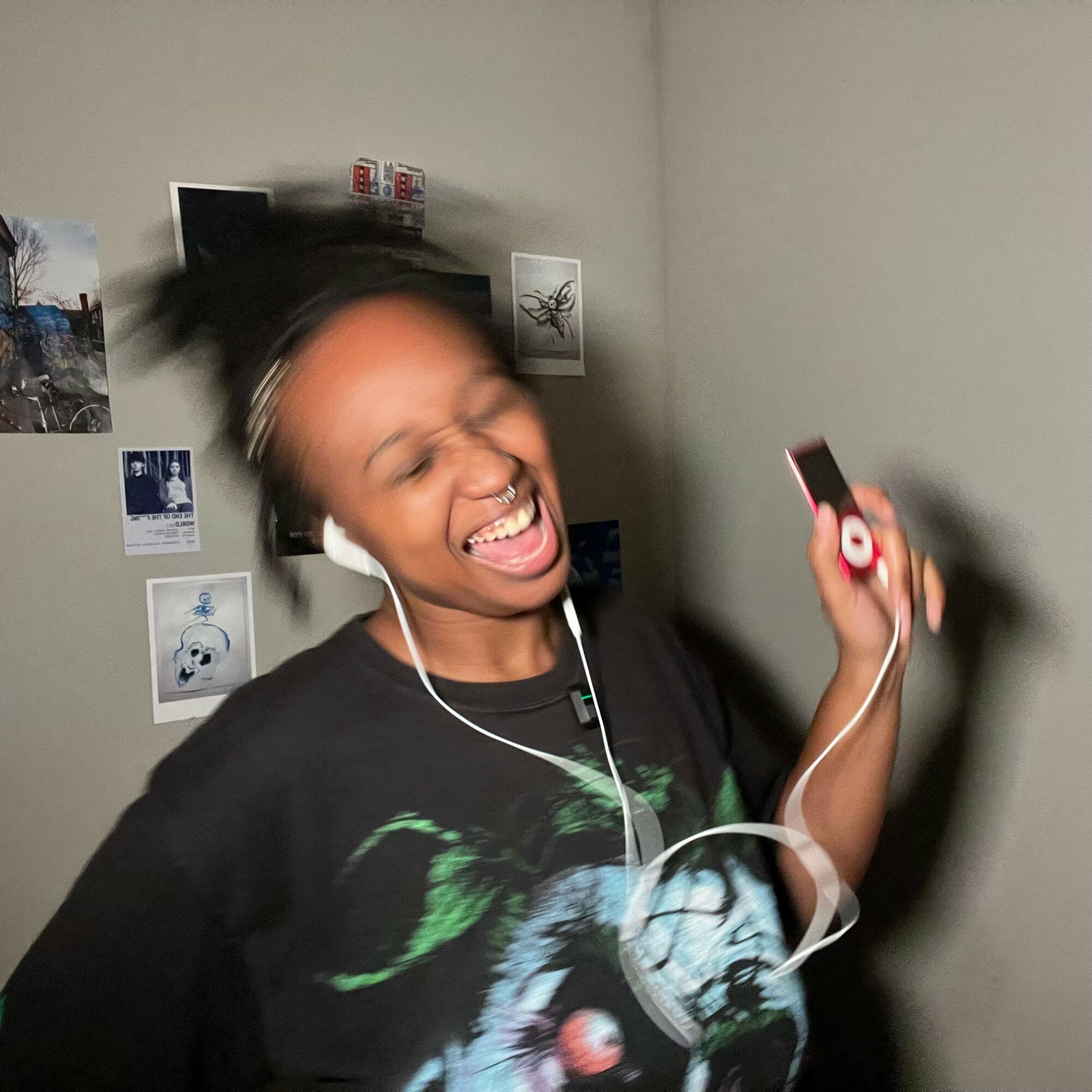 Person smiling and singing while holding a phone, wearing earphones, with a wall decorated with photos behind.