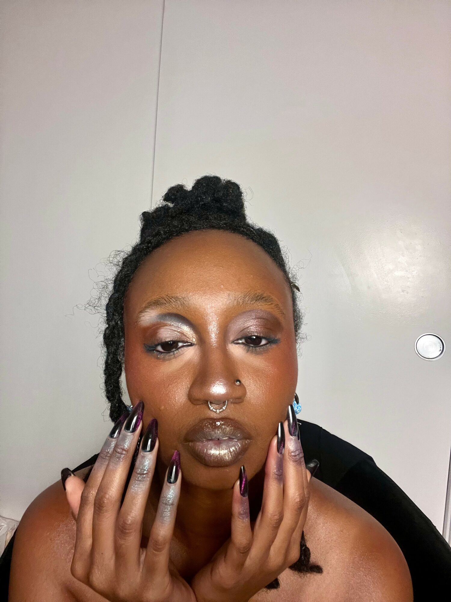 Close-up of a woman with styled hair, makeup, and long nails touching her face, against a plain background.