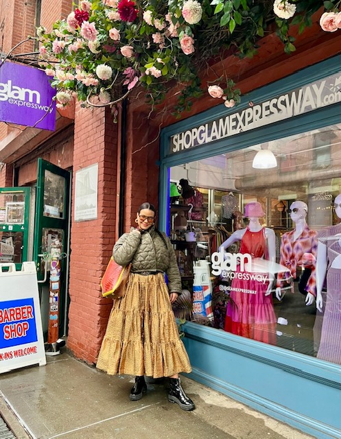 Person standing outside a store with mannequins in the window, pink flowers hanging above, and a sign for Glam Expressway.