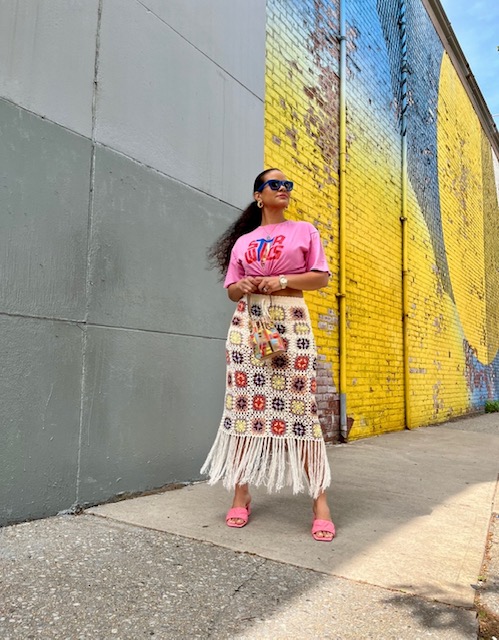Woman standing against a colorful mural wall, wearing sunglasses, pink top, patterned skirt, and pink shoes.