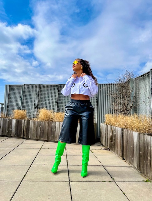 Woman standing outdoors on paved area, wearing sunglasses, cropped top, black pants, and bright green boots, with a blue sky background.