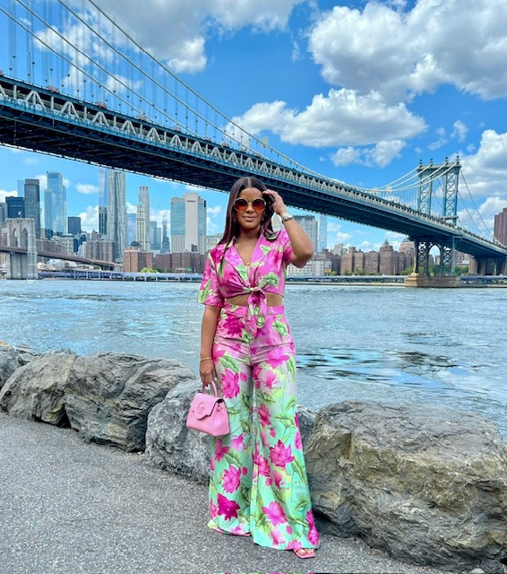 Woman in colorful floral outfit standing on rocks near water with city skyline and bridge in background.