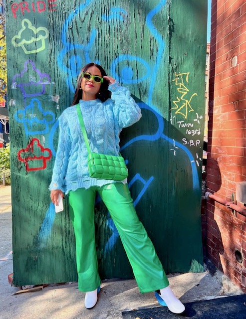Woman in bright green pants and a blue jacket stands in front of a graffiti-covered wall, wearing sunglasses and a green purse.