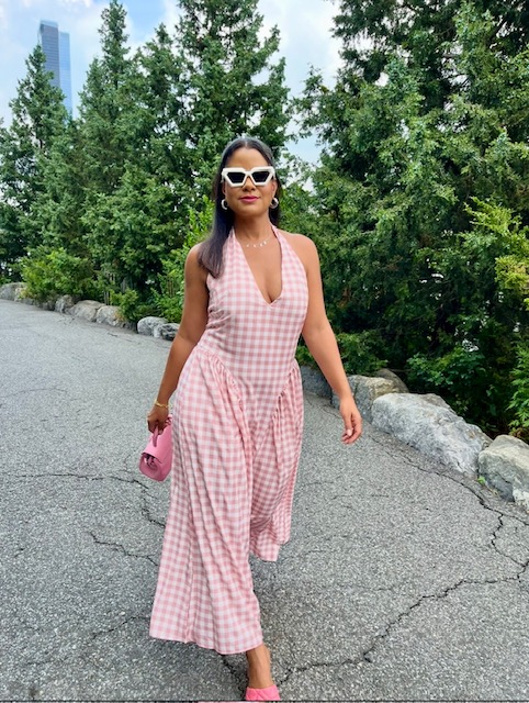 Woman in pink and white checkered dress walking outdoors with trees and rocks in background