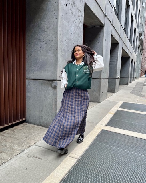 Woman walking on city sidewalk, wearing a green and white jacket and plaid wide-leg pants, smiling, with modern building background.
