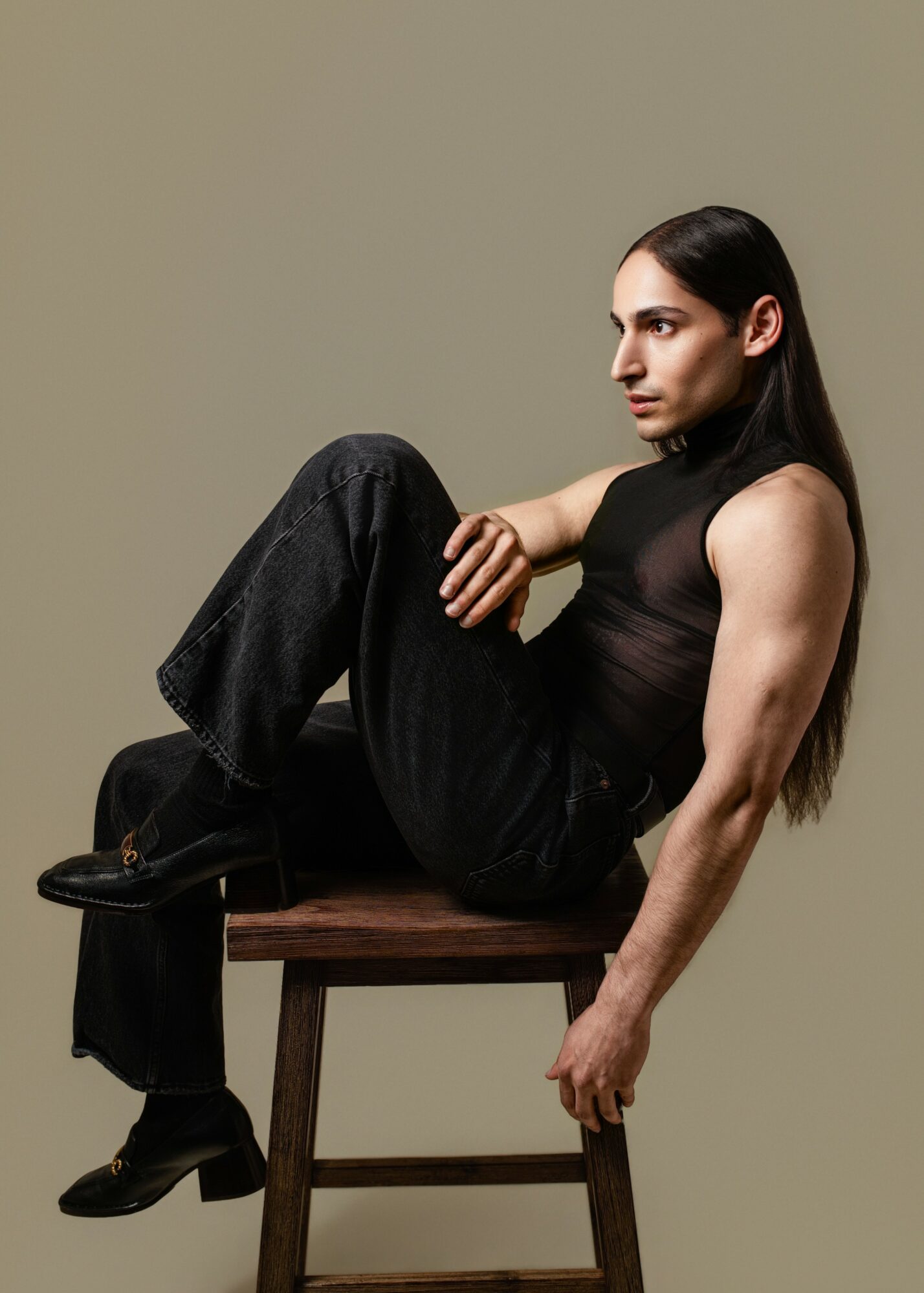 Woman with long dark hair sitting sideways on a wooden chair, wearing a black sleeveless top and black pants, against a plain background.