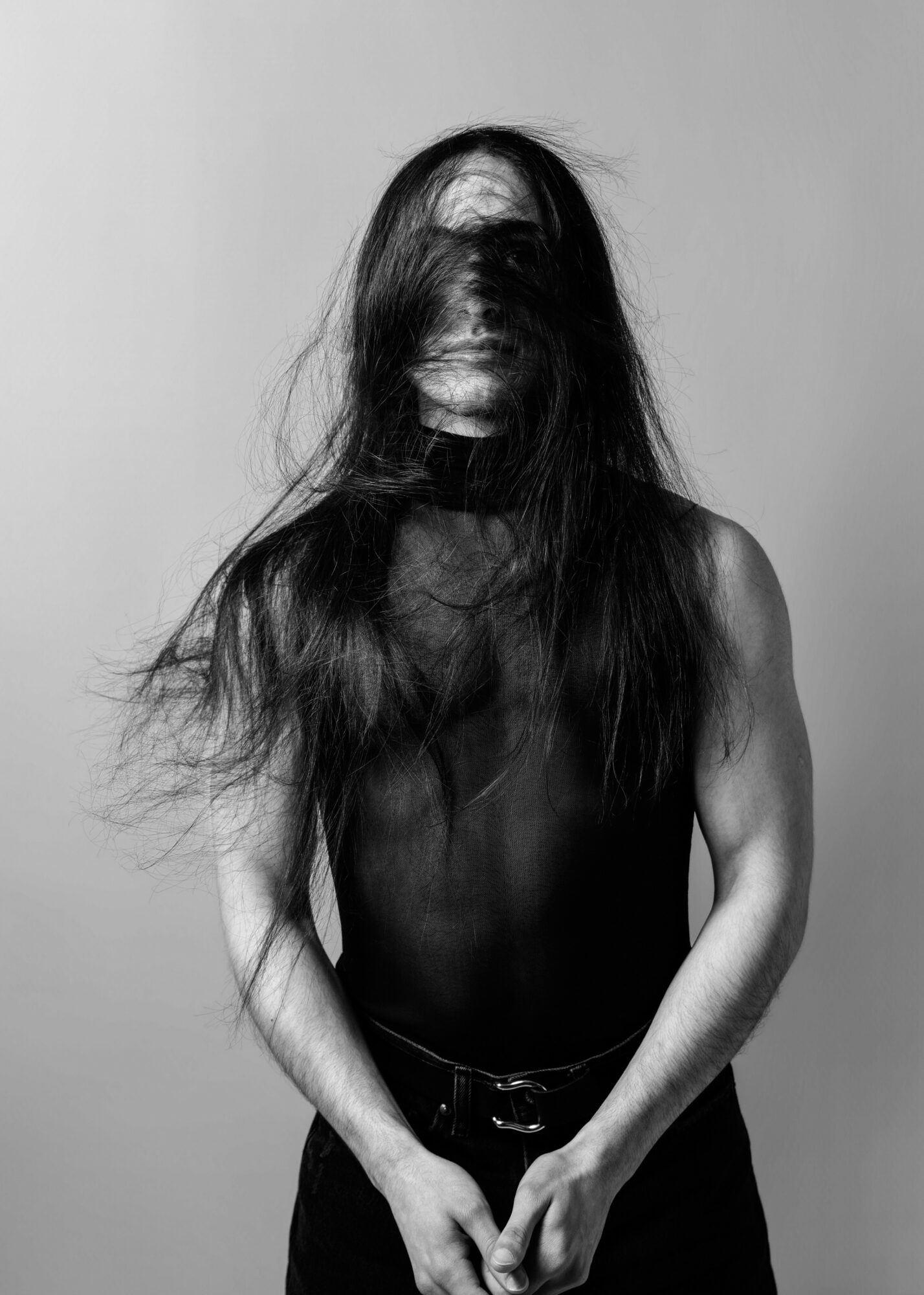 Person with long hair covering face, wearing sleeveless top, standing against plain background.