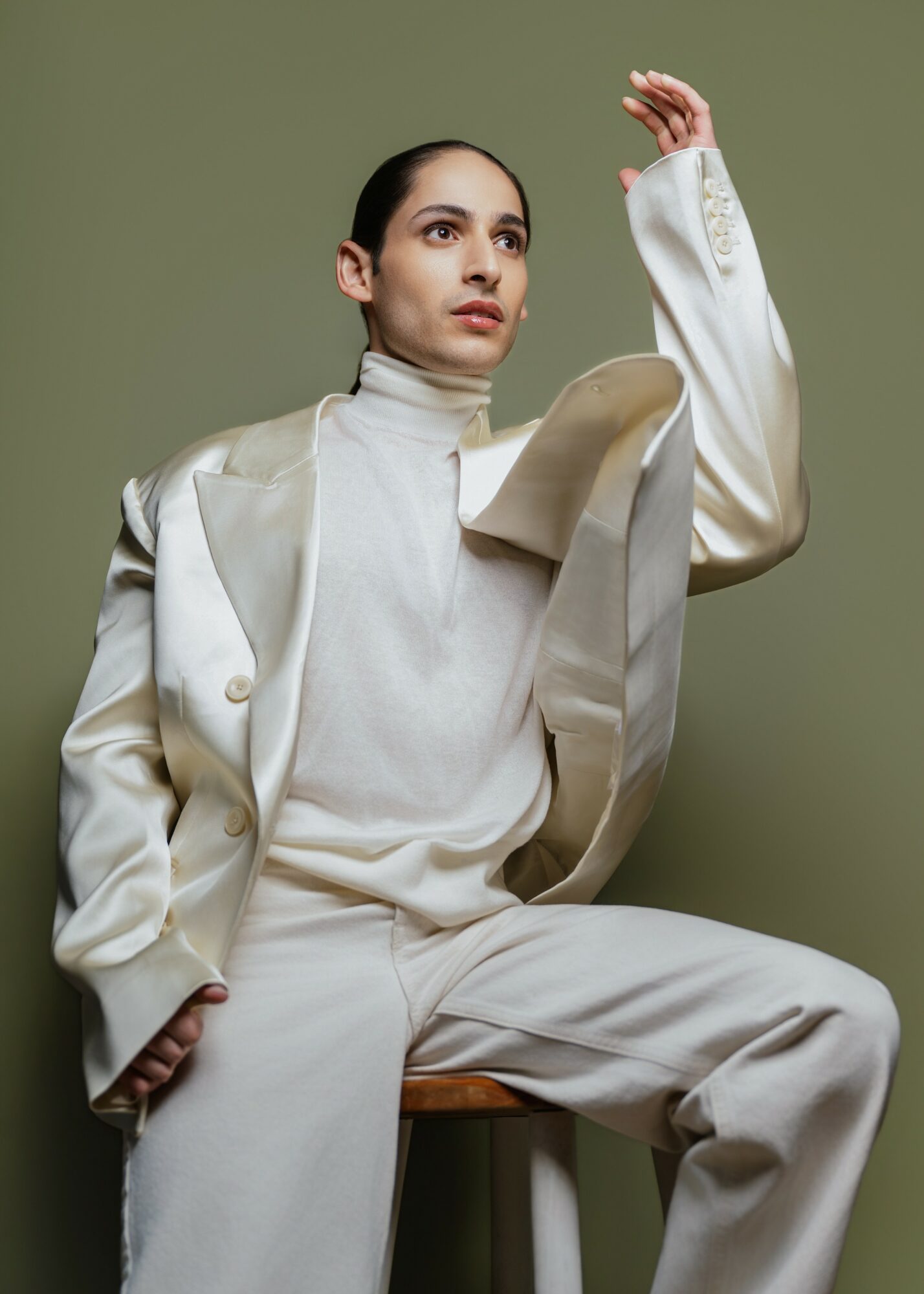 Woman in white outfit sitting on a stool against a green background, looking to the side with her hand raised.