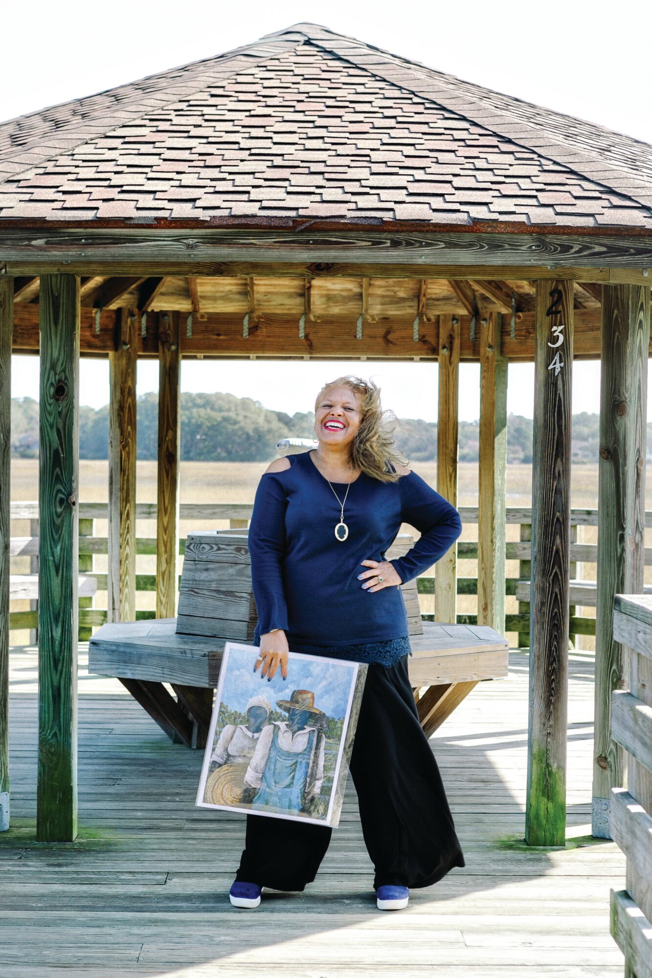Woman standing on a wooden dock holding a framed photo, smiling, with a gazebo and water in background.