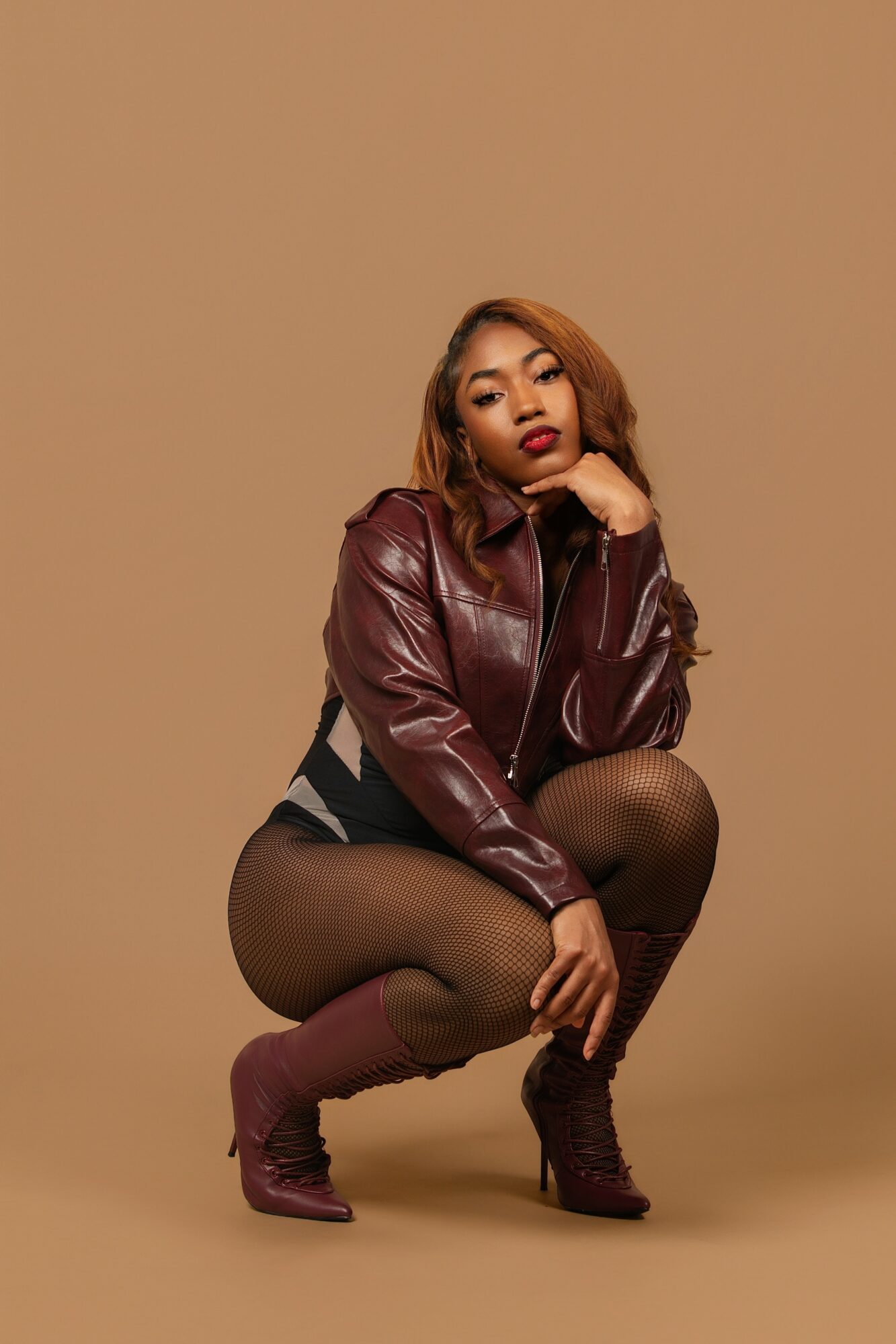 Woman crouching in a leather jacket and high-heeled boots against a plain background.