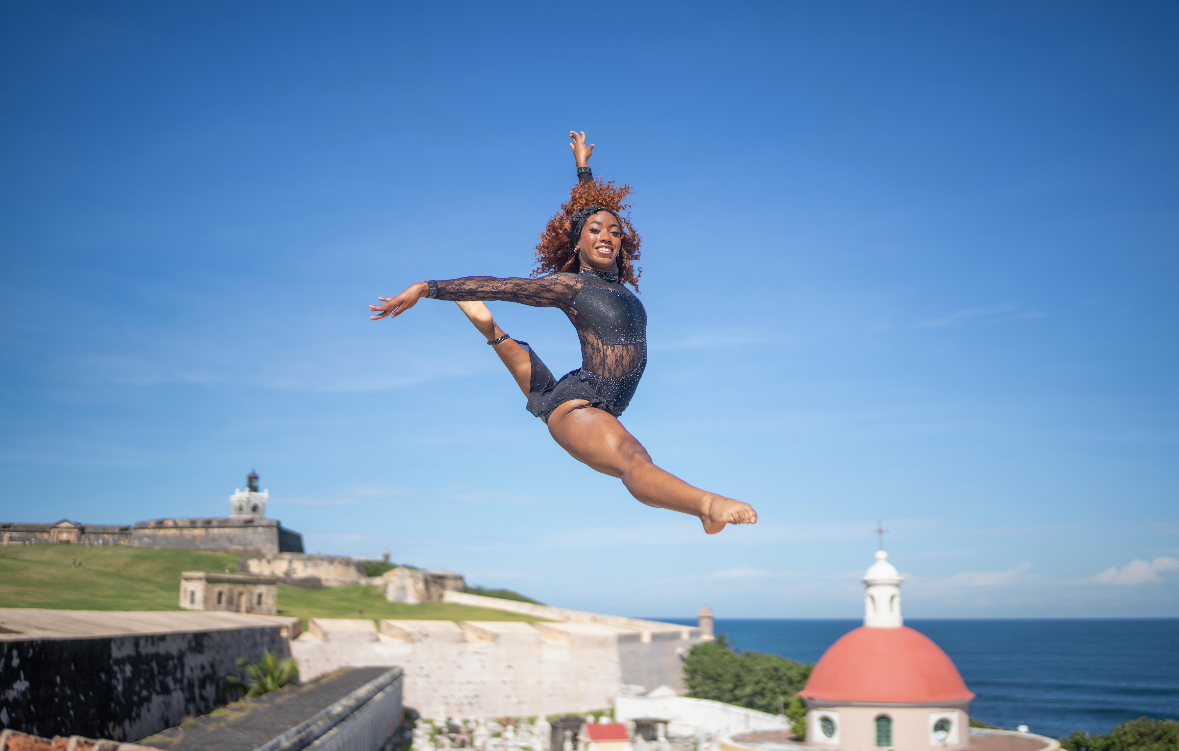 A woman in a leotard jumps in the air against a blue sky with a coastal landscape and lighthouse in the background.