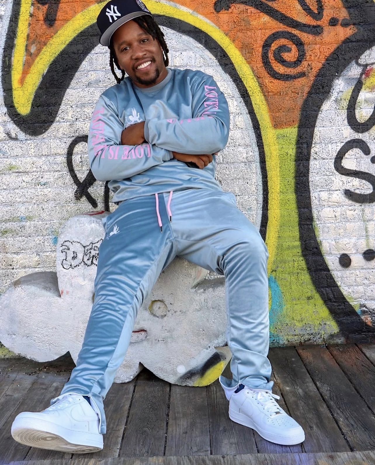 Person sitting on a cloud-shaped bench in front of a colorful graffiti wall, smiling with arms crossed, wearing a light blue tracksuit and a black cap.
