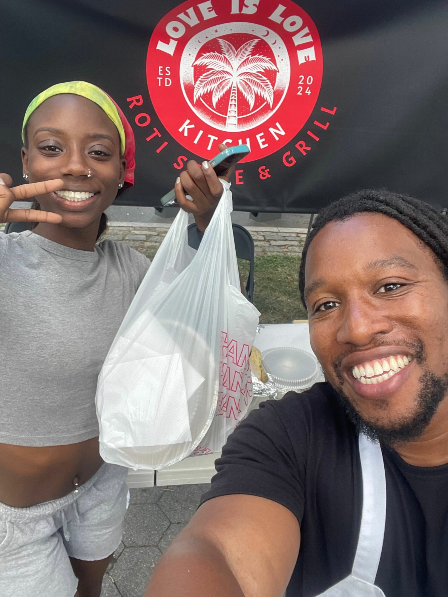 Two smiling people, one holding a plastic bag, outdoors with a red circular sign in the background.
