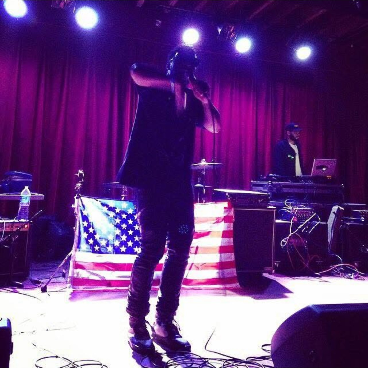 Performer on stage with red curtain, American flag backdrop, and DJ equipment, holding microphone, with stage lights overhead.