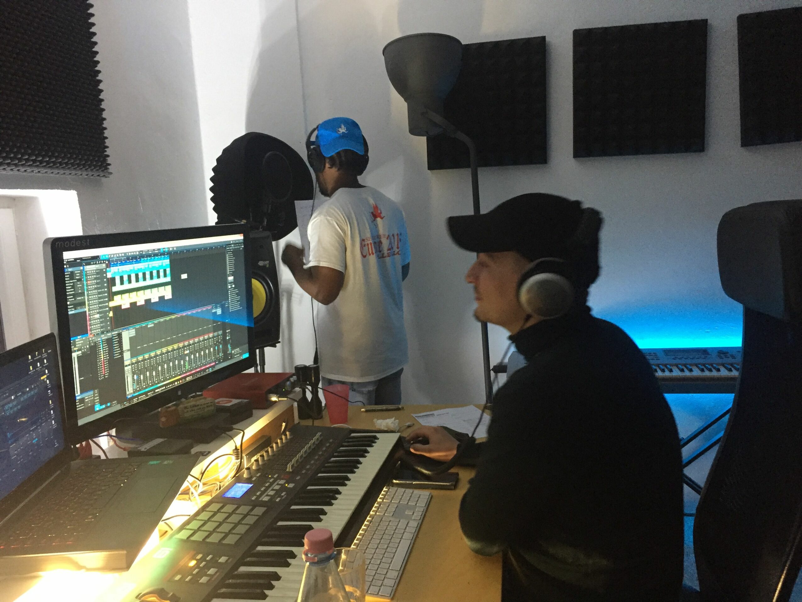 Two people working at a music production setup with a keyboard, monitor, and audio equipment.