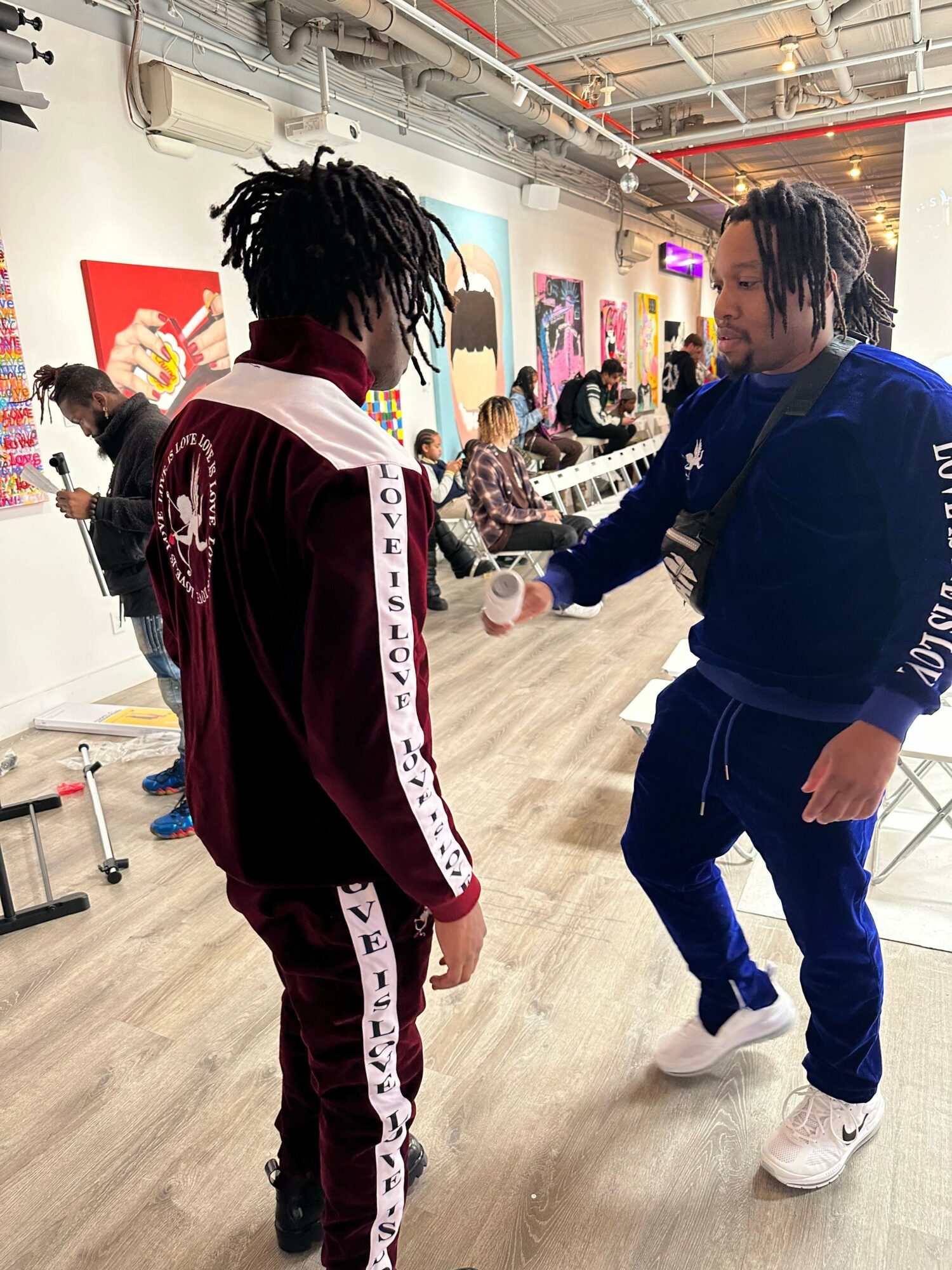 Two people with dreadlocks and casual clothing walk in a brightly lit indoor space with artwork on the walls.