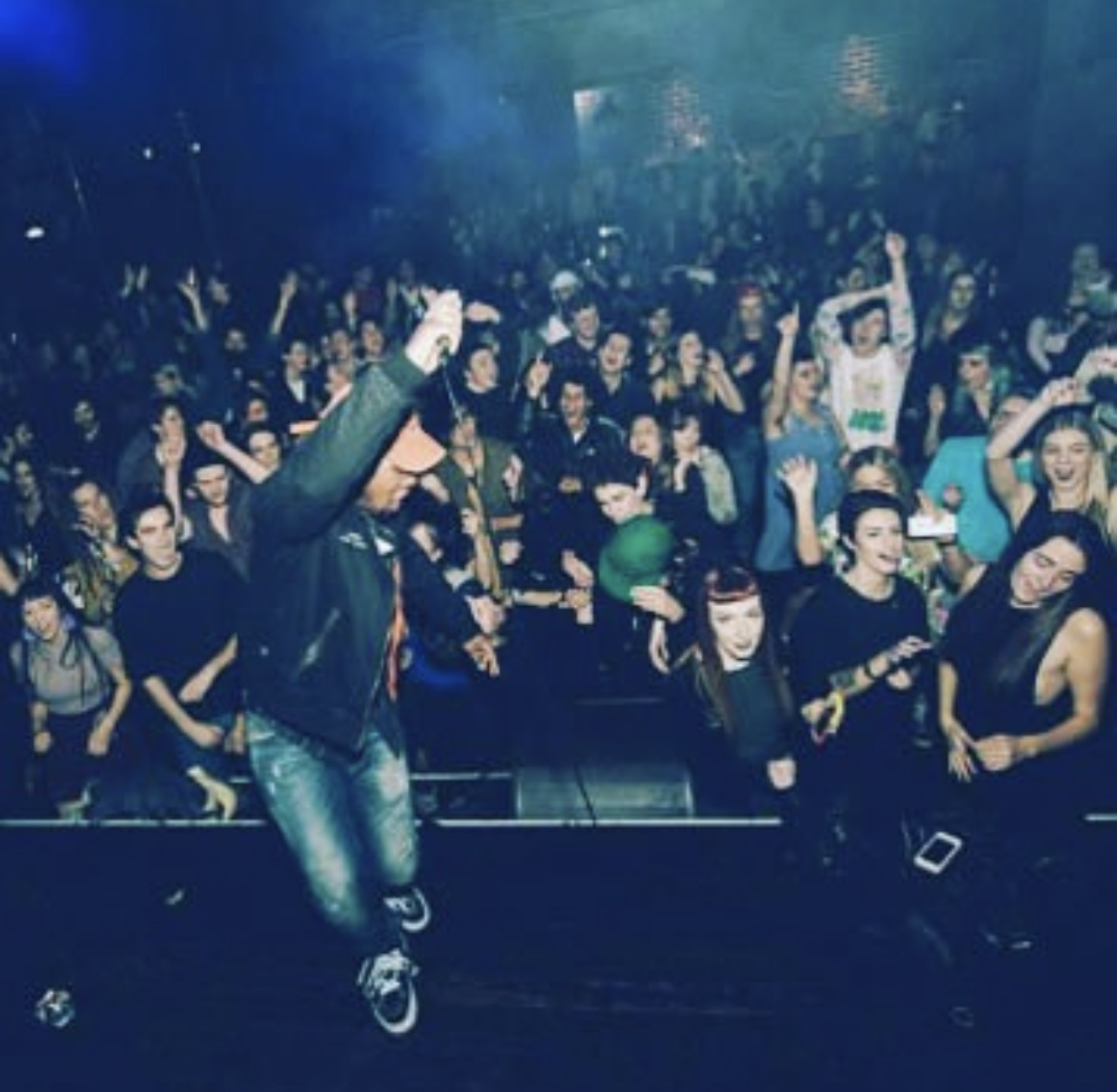 Performer on stage with a crowd of people at a concert or event, some raising hands and holding objects.