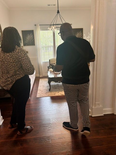 Person standing in a room with hardwood floor, facing away, wearing a black top and patterned pants, with a woman in front.