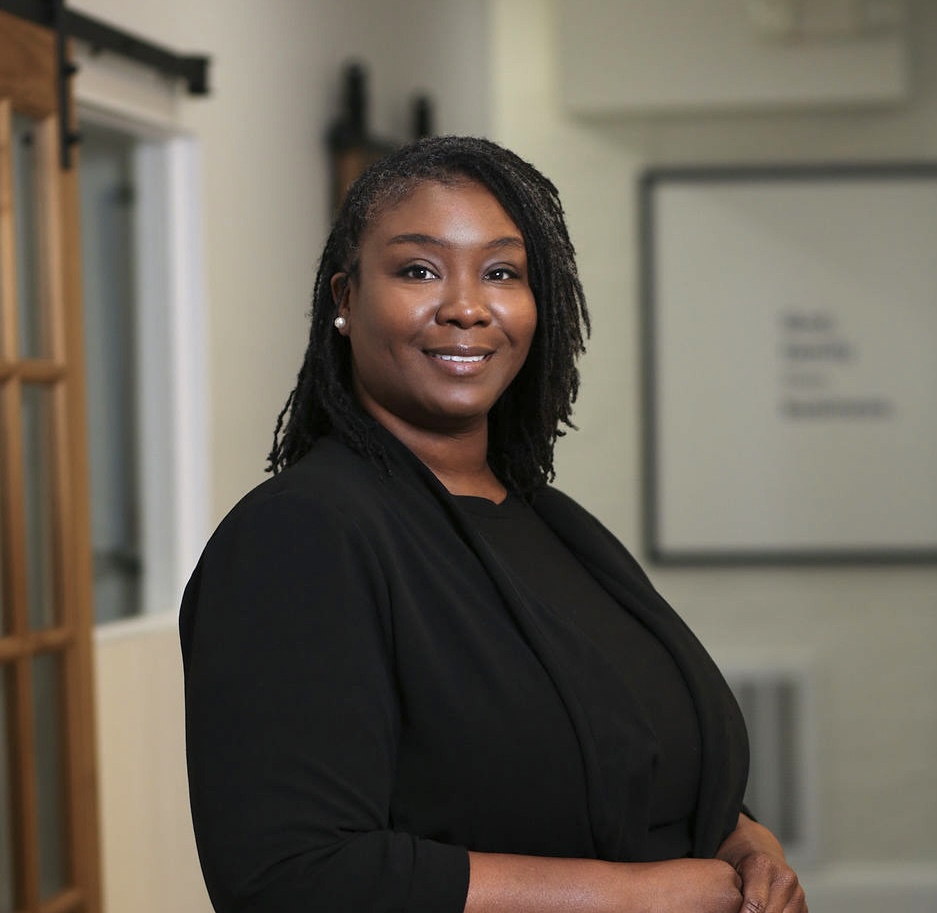 Portrait of a woman with shoulder-length dreadlocks, wearing a black blazer, smiling indoors.