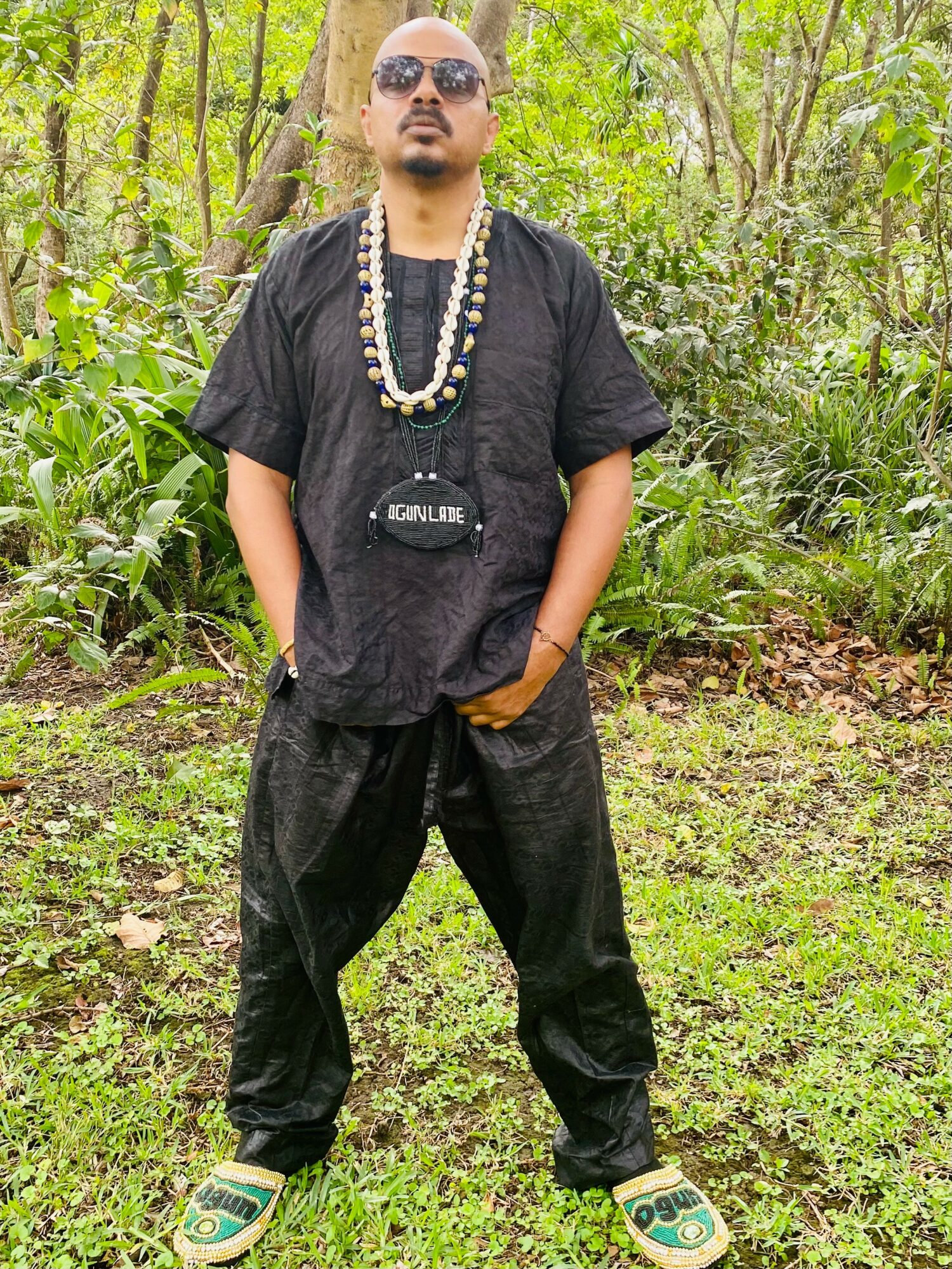 Man standing outdoors in a forested area, wearing sunglasses, a black oversized shirt, baggy black pants, and sandals, with necklaces.