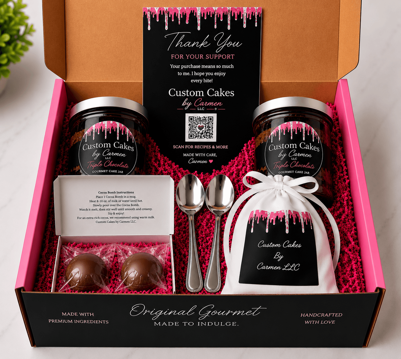 Open gift box with two jars of custom cakes, two spoons, a thank you card, and a wrapped gift, all on pink tissue.