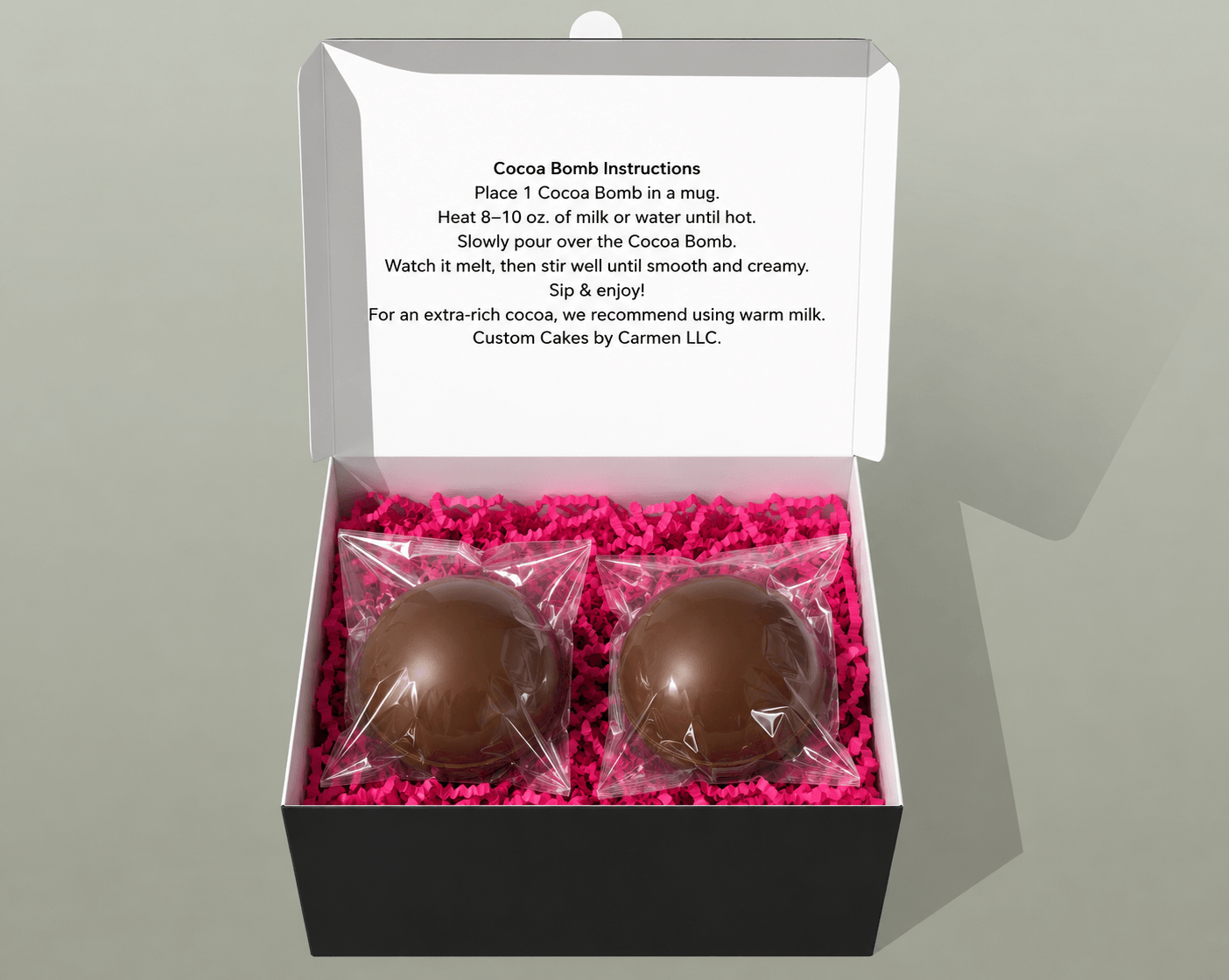 Open box with two chocolate cocoa bombs on pink shredded paper, instruction card inside lid, plain background.