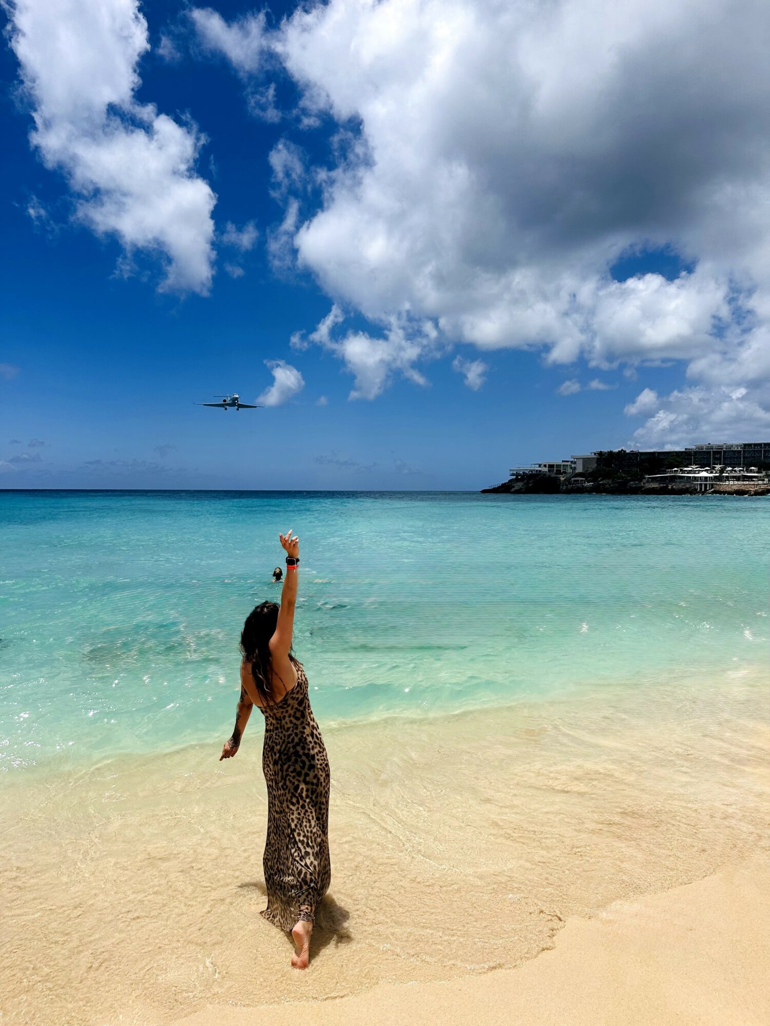 Person standing on sandy beach with arm raised, ocean and sky with clouds in background, airplane flying overhead.