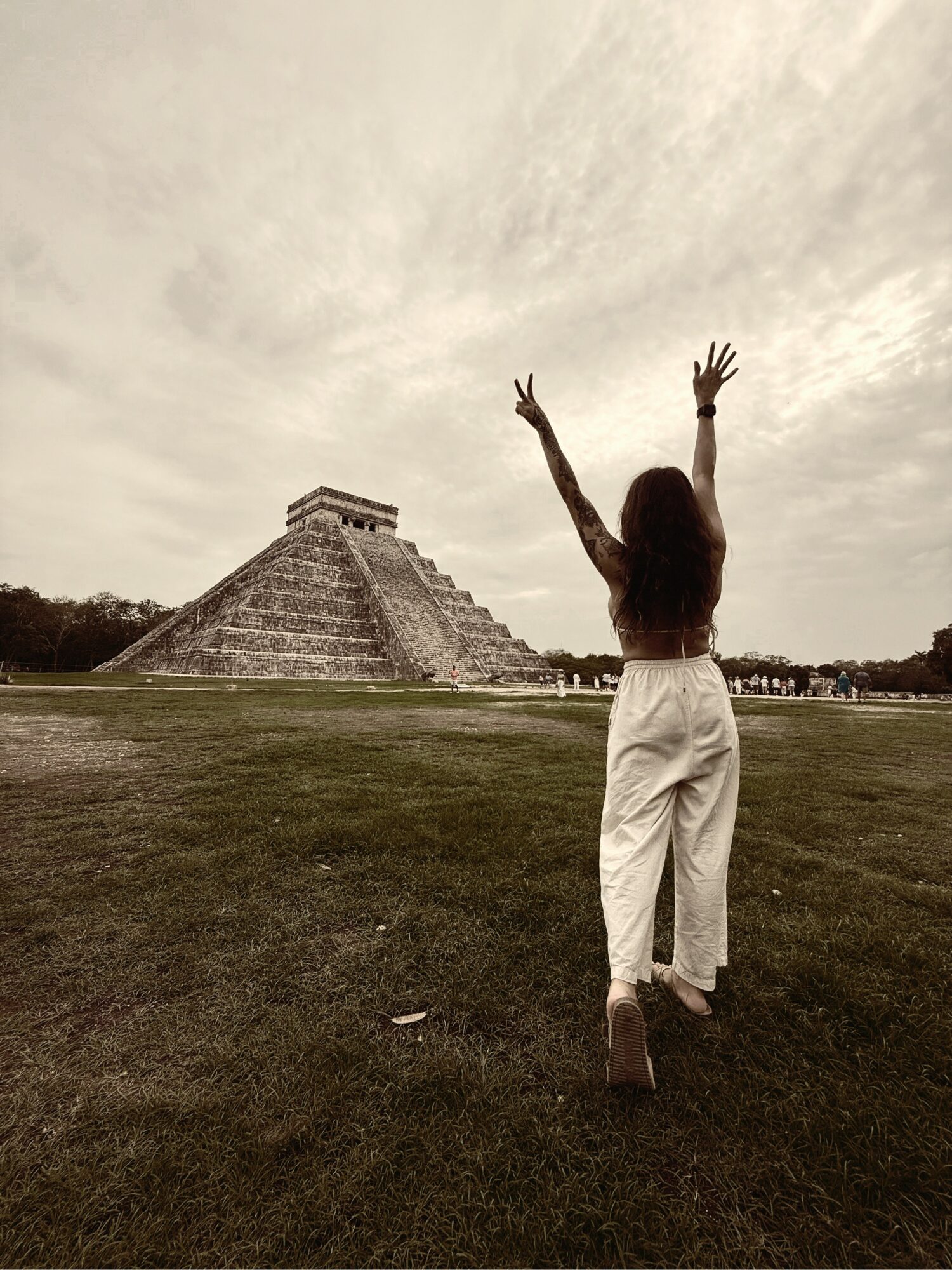 Person with long hair and white pants stands on grass with arms raised, facing ancient pyramid structure under cloudy sky.